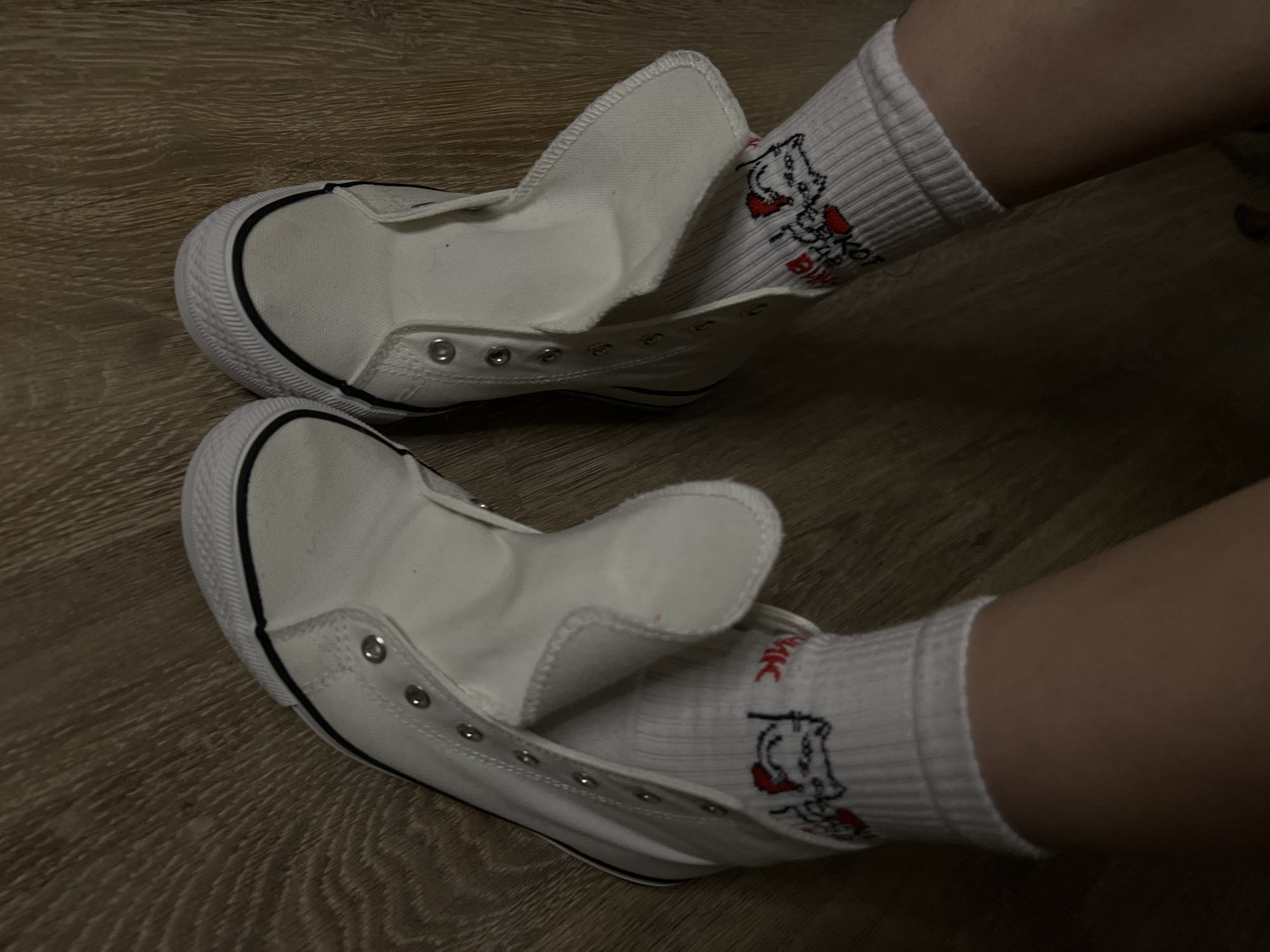 The image shows two feet wearing white socks with cartoon graphics inside white, unlaced canvas sneakers. The sneakers are sitting on a wooden surface with a light brown finish. The background is out of focus. The location is not discernible from the image.