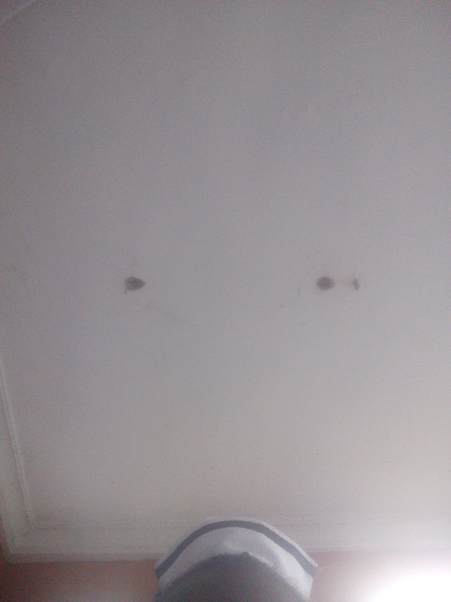 The image shows a ceiling with two dark spots, possibly stains, located approximately one-third of the way from the left side of the frame and two-thirds of the way from the left side of the frame, respectively. The ceiling appears to be white or off-white. In the bottom center of the image, a portion of what appears to be a white hat with grey trim is visible. The background is indistinct but suggests an indoor environment. The image does not provide any information about the city of Kuson, Nigeria, nor does it depict any specific actions or interactions.