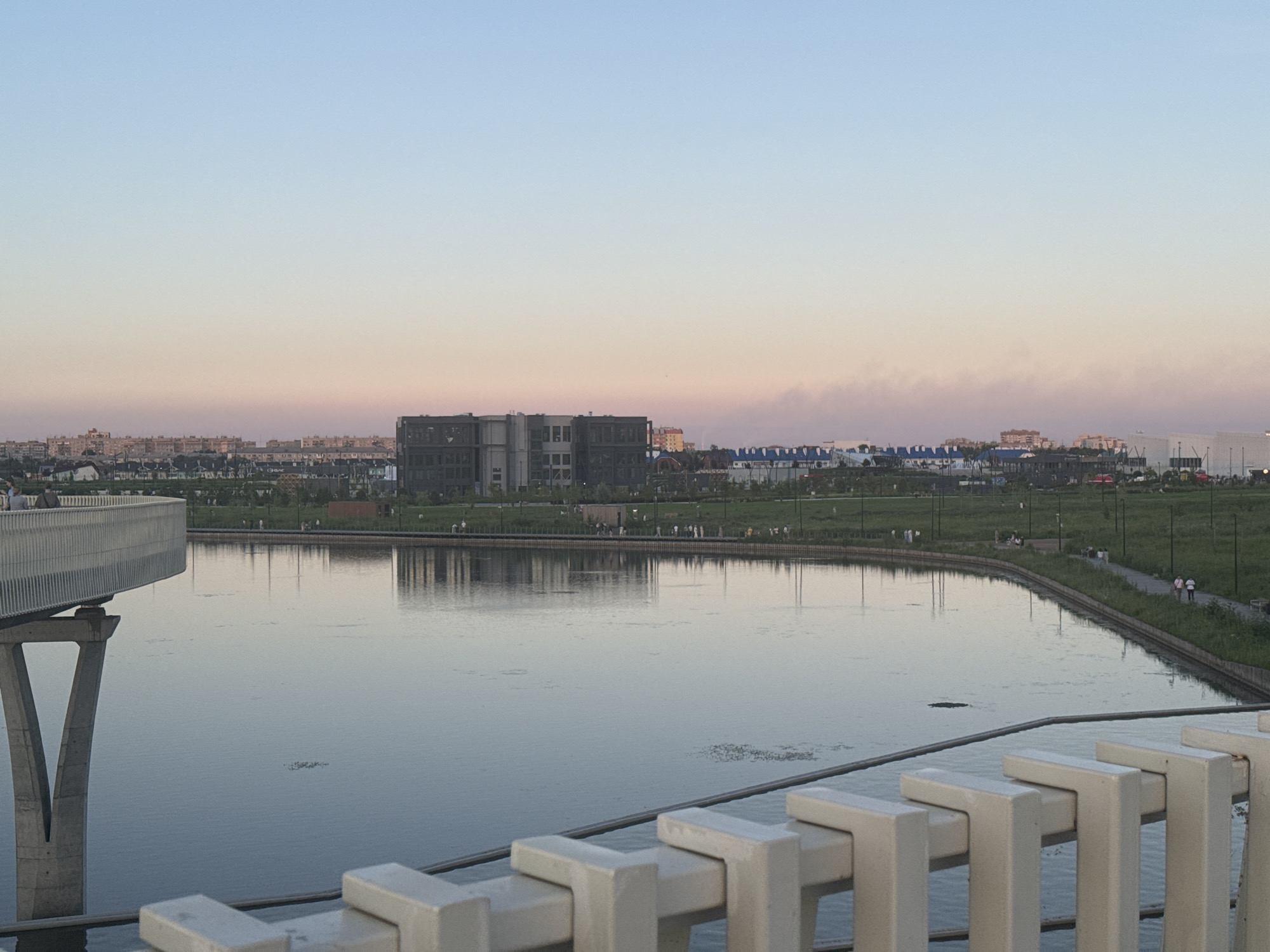 A wide shot captures a tranquil body of water reflecting the soft hues of a sunset, casting a serene mood over the scene. In the foreground, a white, modern railing with geometric detailing guides the viewer's eye towards the middle ground. The railing’s minimalist design contrasts with the organic landscape that unfolds beyond the water.
The middle ground is dominated by a lush green park, dotted with slender lampposts and a paved pathway where several people are seen strolling. The park gently slopes towards the water, creating an inviting public space. Across the park, a striking, multi-story building with a dark, contemporary facade stands as a focal point, its angular architecture creating a strong visual presence. Further in the background, a sprawling urban landscape of low-rise buildings stretches towards the horizon, indicating a populated area.
The sky above transitions from a soft, pale blue at the top to a delicate blend of pink and orange near the horizon, characteristic of a late evening or early morning. A faint haze or smoke drifts across the distant sky, adding a touch of atmospheric depth. The overall impression is one of peaceful coexistence between nature and urban development, with the golden hour lending a sense of calm and beauty to the setting. The location appears to be a public park or waterfront area in Magnitogorsk, Russia, designed for recreation and leisure. No visible text is present in the image.