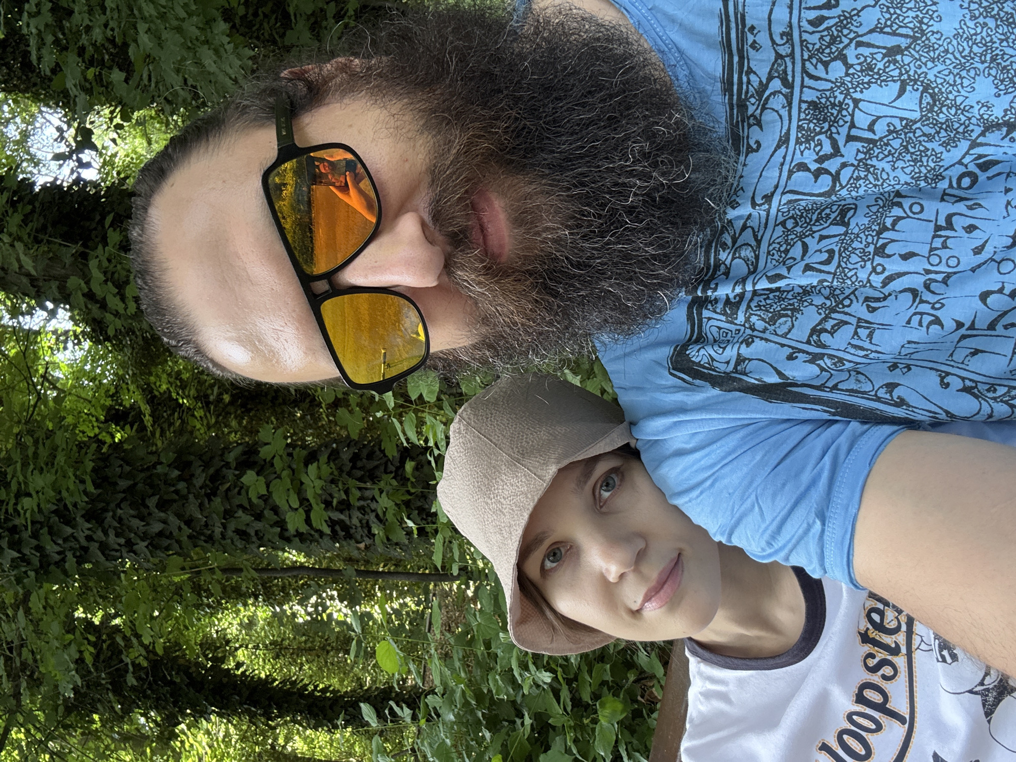 A man and a woman are posing for a selfie outdoors. The man is at the top of the frame, wearing sunglasses that reflect a sunny outdoor scene. He has a large beard and is wearing a blue t-shirt with a black pattern. The woman is positioned below the man. She is wearing a light brown hat and a white t-shirt with black trim and the word "Loopster" printed on it. Green foliage is visible in the background, suggesting they are in a park or wooded area. There is no information available in the image to confirm the location in the city of Tashkent, Uzbekistan.