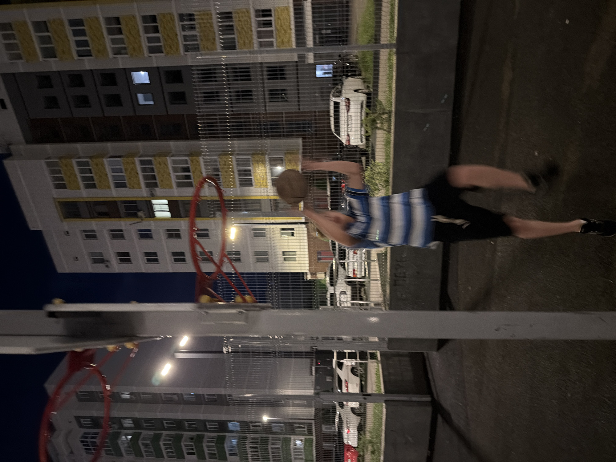 In Izhevsk, Russia, an adolescent male wearing a blue and white striped shirt and black shorts is captured mid-air on a basketball court at night, attempting to shoot a basketball. He is positioned in the foreground, facing away from the camera. The court is dark asphalt, and behind him, a tall, light-colored apartment building with yellow accents and numerous illuminated windows stands under a dark sky. A chain-link fence separates the basketball court from a parking area where several white cars are parked. The basketball hoop, with its red rim and netting, is visible in the upper left corner of the frame. The scene suggests a casual recreational activity taking place in a residential area.