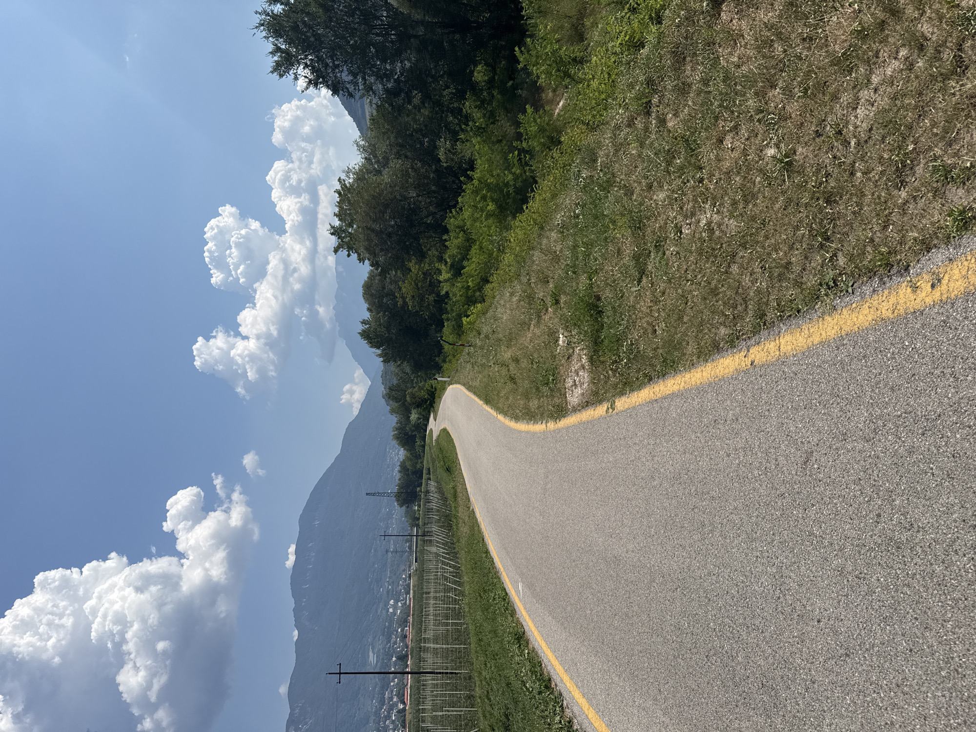 The image depicts a paved, winding road with a yellow line marking its edge, likely a bike path or a quiet road, cutting through a rural landscape. On the left side of the road, a field with numerous vertical poles and wires, possibly a vineyard or orchard, stretches into the distance. Beyond the field, a town with buildings is visible, nestled against rolling hills. In the background, majestic mountains rise under a bright blue sky dotted with fluffy white clouds. The right side of the road is bordered by lush green vegetation, with trees and bushes forming a dense natural barrier. The scene suggests a pleasant, sunny day in a scenic, possibly mountainous region. This road is likely located in the outskirts of Trento, Italy, given the distinctive mountain landscape and the presence of vineyards, common in the Trentino-Alto Adige region.