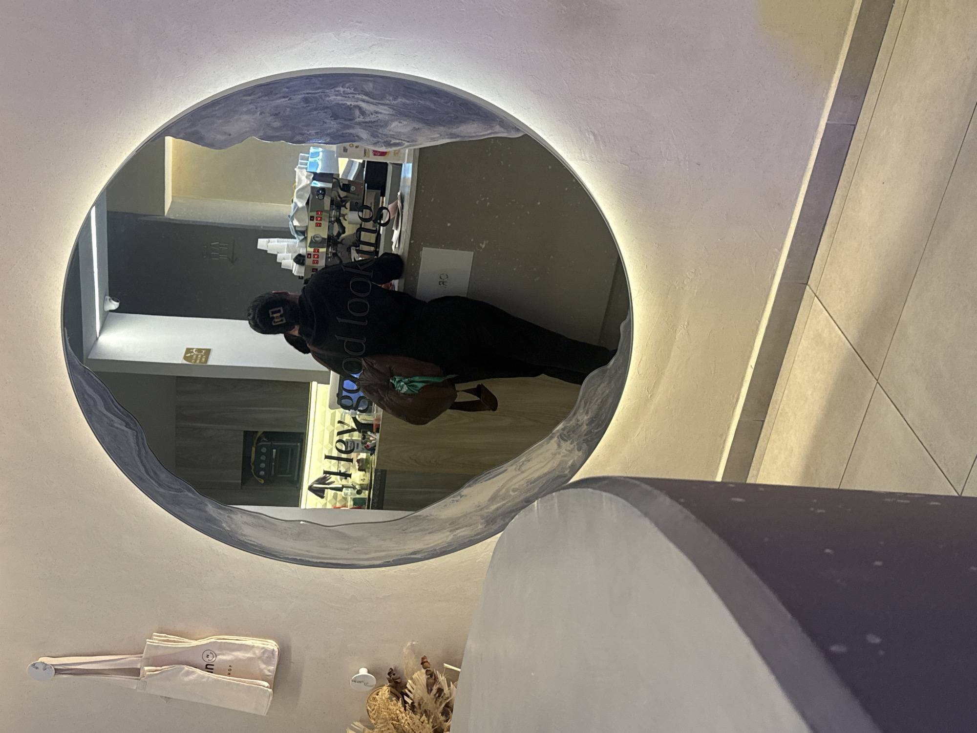 A mirror reflects a person in black clothing standing at a counter in a cafe, likely in San Bernardino Tlaxcalancingo, Mexico. The counter has a beverage machine and drink containers. The reflection also shows the cafe's interior, including a menu and a display case. A tote bag hangs near the mirror, and part of a gray cylindrical structure is visible in the foreground. The scene is lit with ambient light.