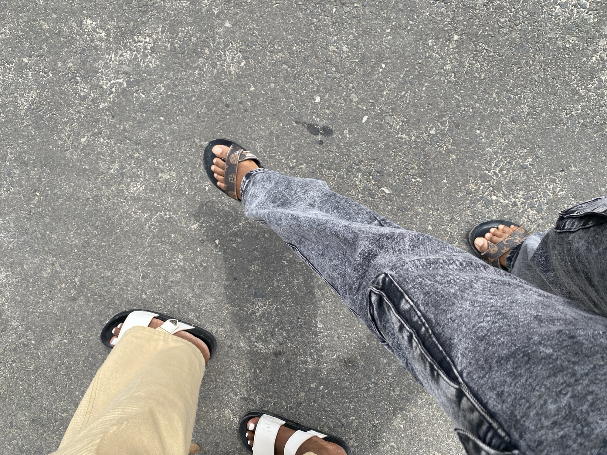 The image depicts an overhead view of the lower legs and feet of two individuals on a coarse-grained asphalt surface in Dar es-Salaam, Tanzania.
One individual, positioned in the upper left, wears beige-colored pants and black-soled sandals with two broad white straps. The toes of this individual, visible through the sandals, are painted with white nail polish. Partially visible are the left foot and the anterior portion of the right foot.
The second individual, occupying the lower right and center of the frame, wears acid-wash grey denim cargo pants. This person is wearing black-soled sandals featuring brown straps patterned with a repeating monogram design. Both feet of this individual are visible, with the left leg extended forward.
The environment consists of dark grey asphalt pavement, showing various light-colored flecks and several small, irregular dark stains, possibly from liquid. The overall impression is an outdoor urban or semi-urban setting where pedestrians are present. No other subjects or objects are discernible beyond the individuals' lower extremities and the ground. The individuals appear to be in motion or standing on the paved surface.