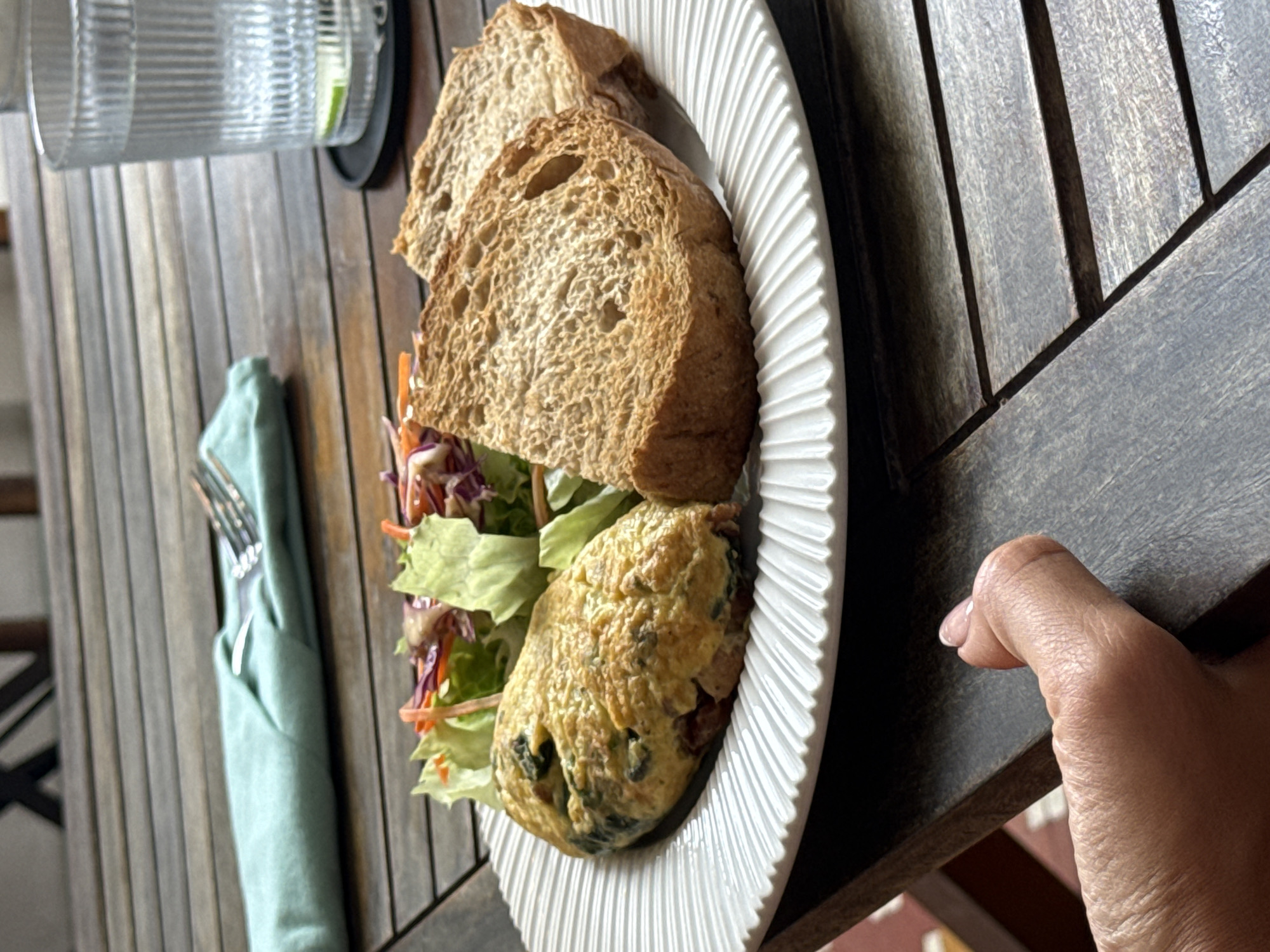 The image shows a plate of food on a wooden table. The plate contains two slices of whole wheat bread, a scoop of salad, and an omelet. A glass of water and a wrapped utensil are also visible on the table. A person's hand is present in the image, near the corner of the table. The image was likely taken in Đà Nẵng, Vietnam.