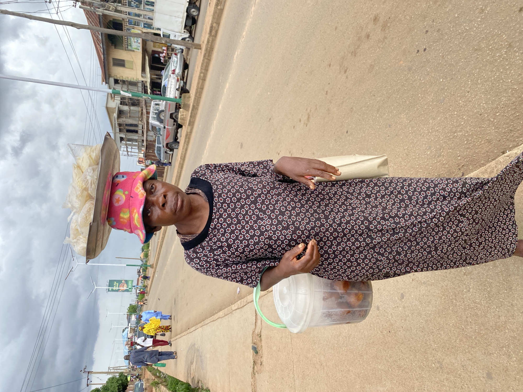 A medium shot captures a woman balancing a tray of packaged goods on her head while walking on a paved sidewalk. She is wearing a dark, floral-patterned dress and a brightly colored, multi-patterned bucket hat. The woman's right hand holds a beige cloth bag. A clear plastic bucket with a green handle, containing food items, hangs from her left arm. The background shows a street scene with vehicles, buildings, and overhead power lines, suggesting an urban environment. The sky is overcast with dark clouds. The scene is likely located in Bukuru, Nigeria, based on the provided information.