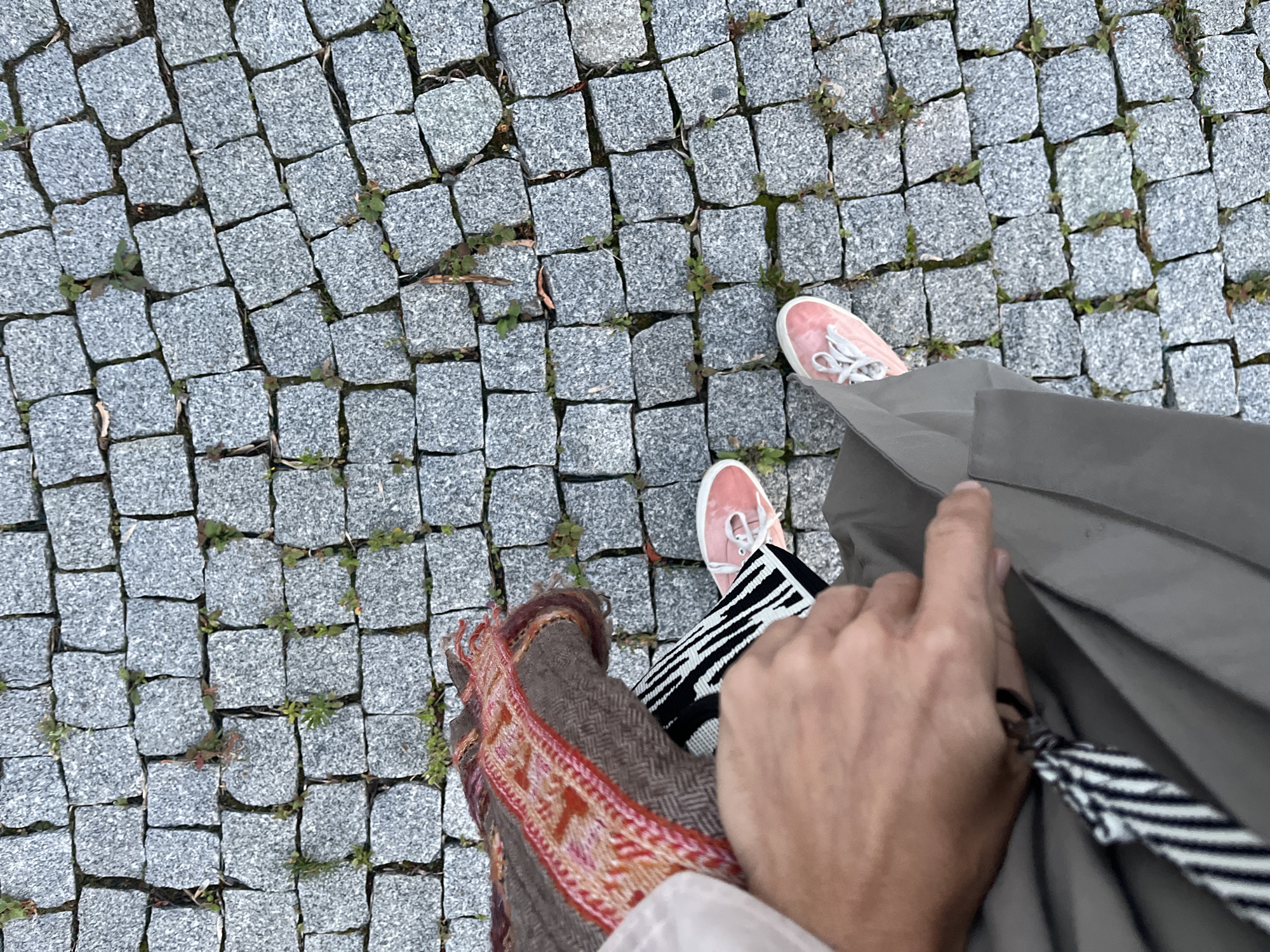 This image captures a top-down view of two people standing on a cobblestone path in Moscow, Russia. One person is wearing pink sneakers and a gray coat, while the other is wearing similar pink sneakers and a brown scarf with a colorful woven pattern. The cobblestone path is uneven, with small green plants growing in the cracks between the stones, suggesting an outdoor setting. The lighting is diffuse, indicating it might be an overcast day or late afternoon. The activity appears to be casual, perhaps walking or pausing during an outing. The overall mood is neutral, with no strong emotional cues. There is no visible text in the image.
