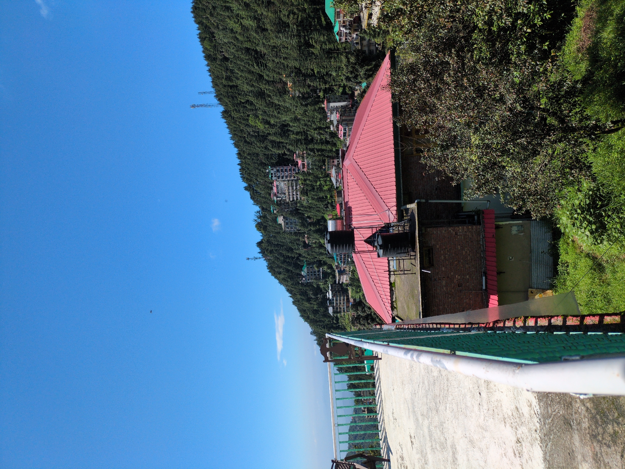 In Bhagsu, India, a clear blue sky dominates the upper portion of the frame, with a faint wisp of a cloud visible on the horizon. Below, a lush green hillside is densely covered with evergreen trees, interspersed with buildings featuring red and green roofs. The buildings appear to be residences or guesthouses, clustered on the slopes. In the foreground, a concrete patio area leads to a railing with green metal mesh and white metal bars, offering a view over the valley. Two empty chairs are positioned on the patio. A red-roofed building with two black water tanks on its rooftop is partially visible in the mid-ground, adjacent to other brick and concrete structures. The overall scene depicts a tranquil mountain town setting on a sunny day.