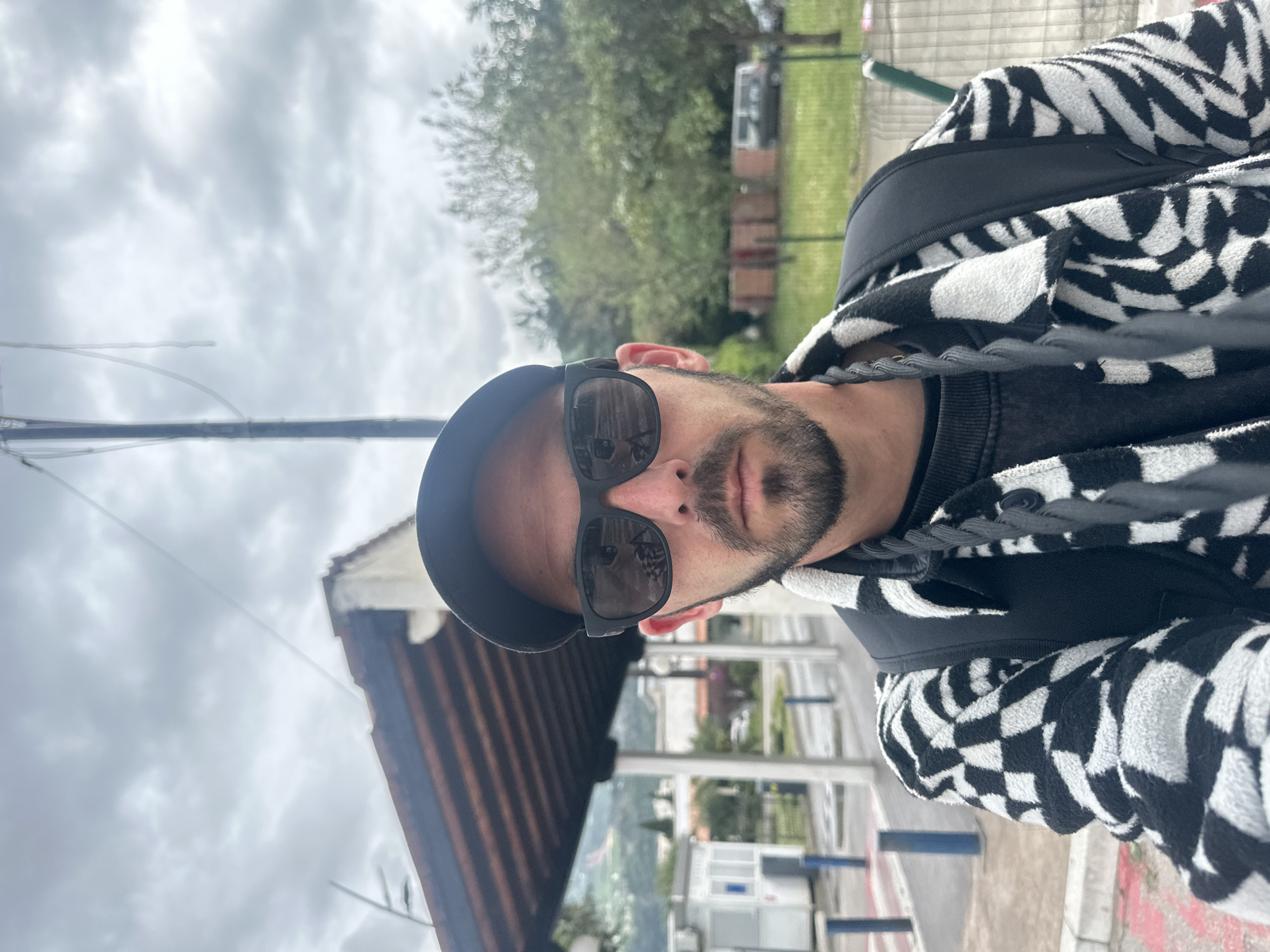 This is a selfie taken by a man outdoors on an overcast day, likely in Budva, Montenegro. The man is centered in the foreground, looking directly into the camera. He wears a black baseball cap, dark sunglasses that reflect parts of the environment, a dark long-sleeved shirt, and a distinctive black and white patterned hoodie or jacket with a geometric or checkerboard design. He has a beard and mustache, and his expression is neutral, with his eyes obscured by the sunglasses.
The background features a cloudy, grey sky dominating the upper half of the image, indicating overcast weather during the daytime. To the man's left, a slender utility pole rises against the sky. To his right and extending into the mid-ground, green trees and foliage are visible. Further in the distance, various buildings with visible roof structures are present, suggesting a town or residential area. The perspective implies an elevated vantage point overlooking the landscape. No visible text is present in the image.