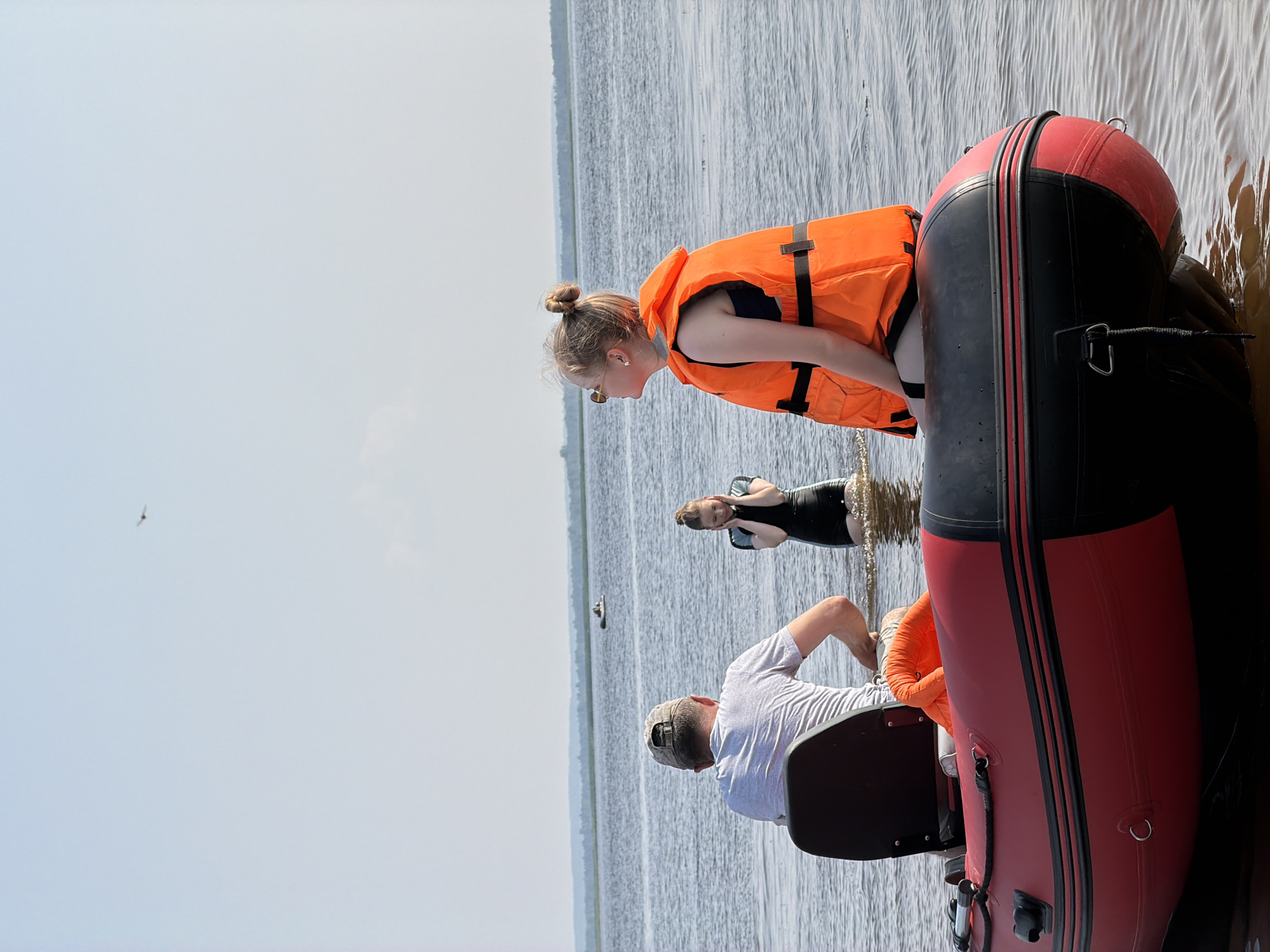 In Krasnyi Bor, Russia, on a bright, clear day, a red and black inflatable boat floats on a calm body of water. A man wearing a grey t-shirt and a baseball cap sits in the boat, facing away from the camera, with his arms submerged in the water. Seated in front of him, also facing away from the camera, is a young woman wearing a bright orange life vest over a dark blue swimsuit. Her hair is pulled up in a bun, and she wears sunglasses. Another young woman, in a black and teal wetsuit, stands in the water to the left of the boat, with her hands covering her ears. In the distance, the shoreline is visible with a line of trees, and another small boat with a person in it is on the water. A bird is seen flying in the hazy sky.
