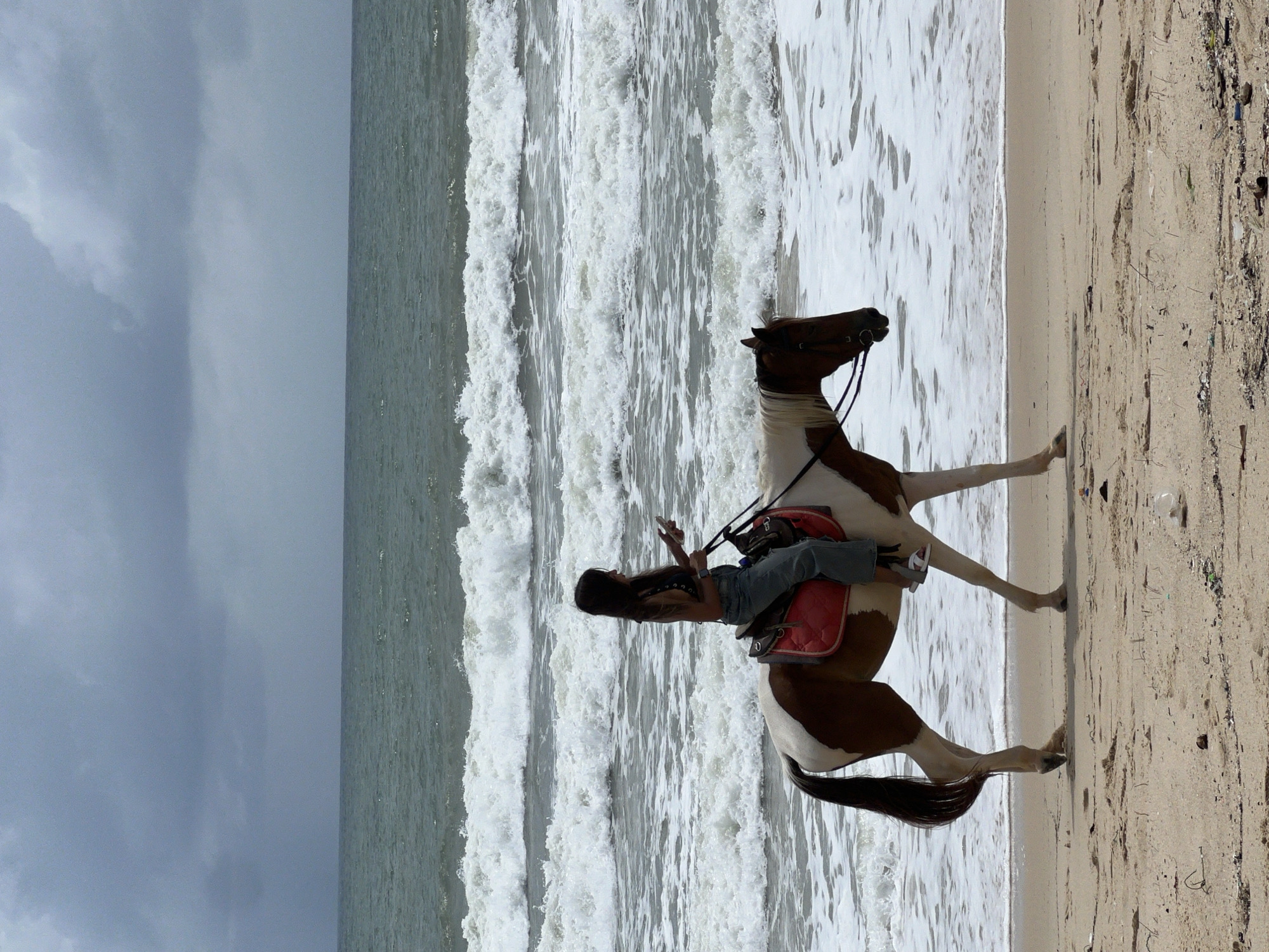 A woman rides a brown and white horse along a sandy beach, with the ocean and waves in the background. The sky is cloudy. The woman is wearing jeans and a dark top and is looking at her phone. The horse is wearing a red saddle pad and a bridle. The scene takes place in Choeng Thale, Thailand.