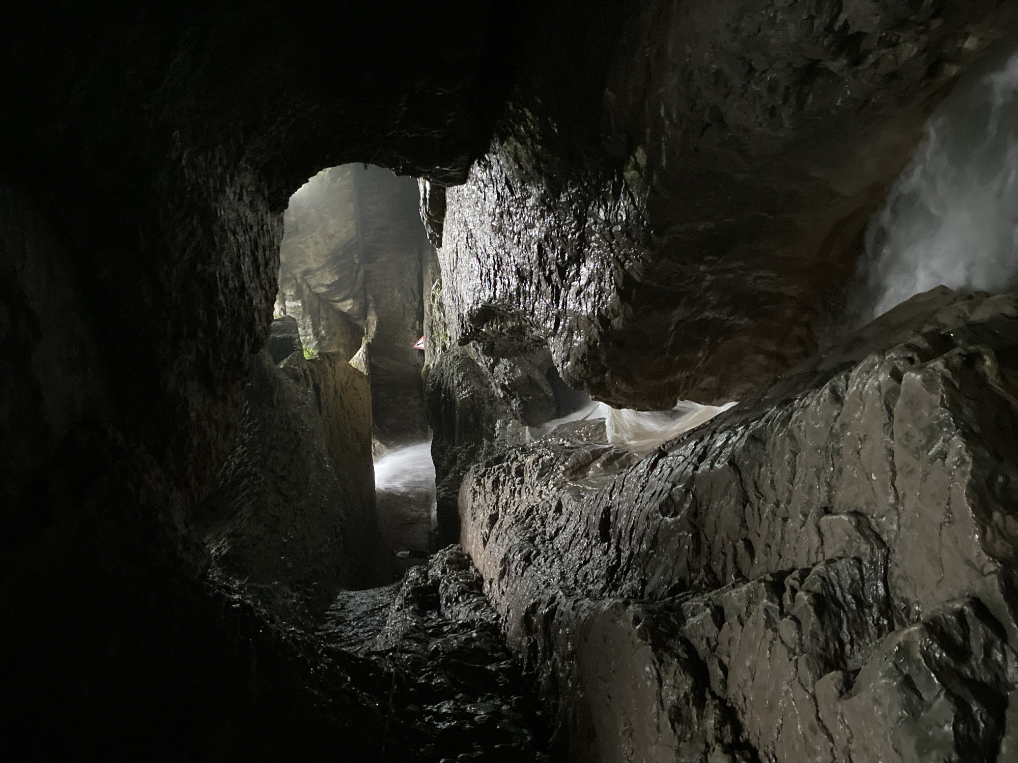 The image depicts the interior of a cave with a narrow passage carved by a stream. The walls of the cave are rough and wet, with visible layers of rock. Water flows rapidly through the passage, creating a frothy white appearance. In the background, a faint light illuminates the scene, suggesting an opening to the outside. There are two small figures visible in the distance, one standing on a ledge and another near a waterfall. The environment is damp and appears to be a natural geological formation. This scene is likely located in the vicinity of the Salta village in Russia, known for its numerous canyons and waterfalls.