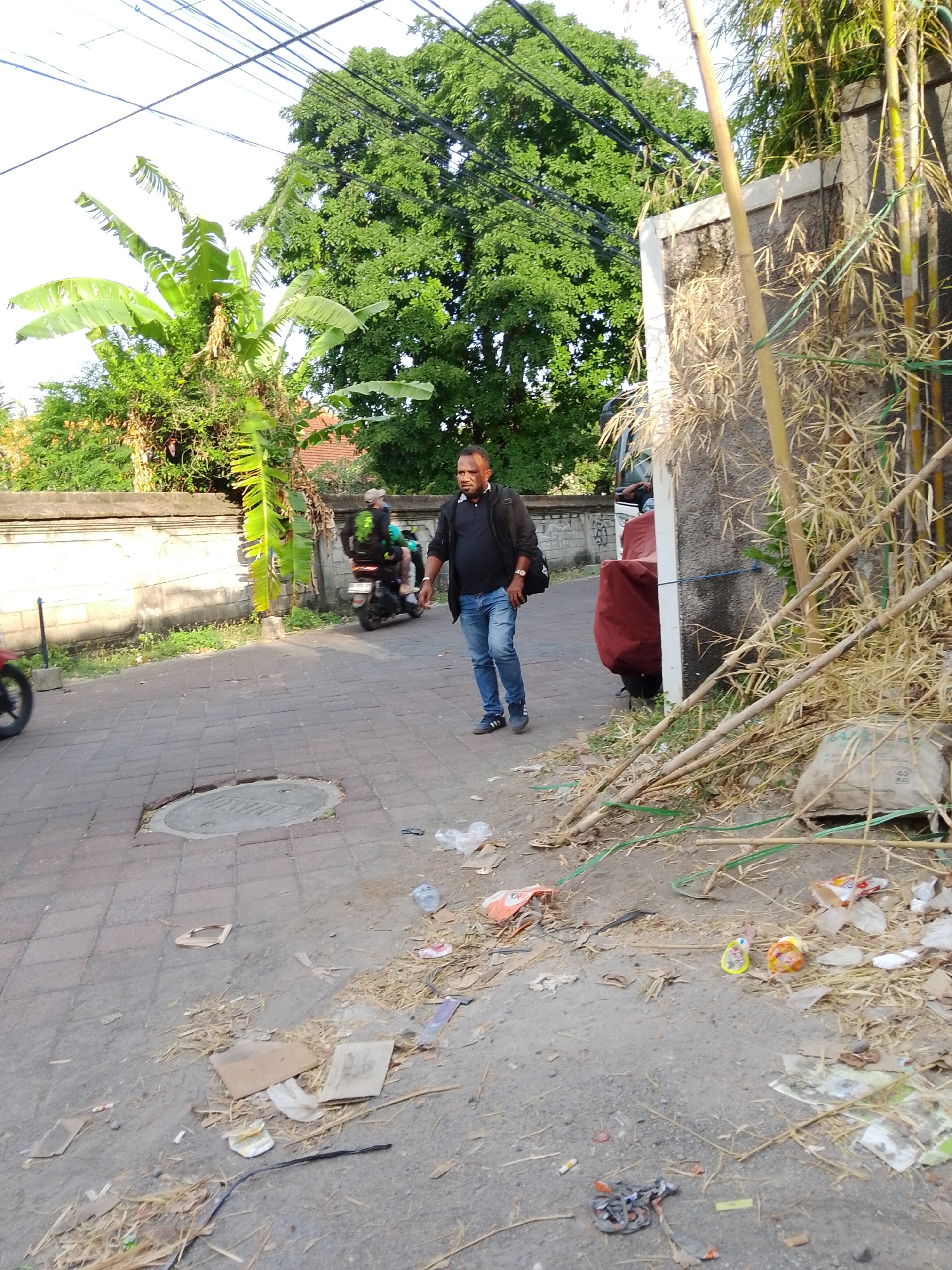 The image shows a street scene in Kuta, Indonesia. A man in a black jacket and blue jeans is walking on a paved walkway. In the background, a motorcycle is visible. The area appears to be somewhat cluttered with debris and vegetation. The overall impression is that of a typical street in a tropical urban environment.