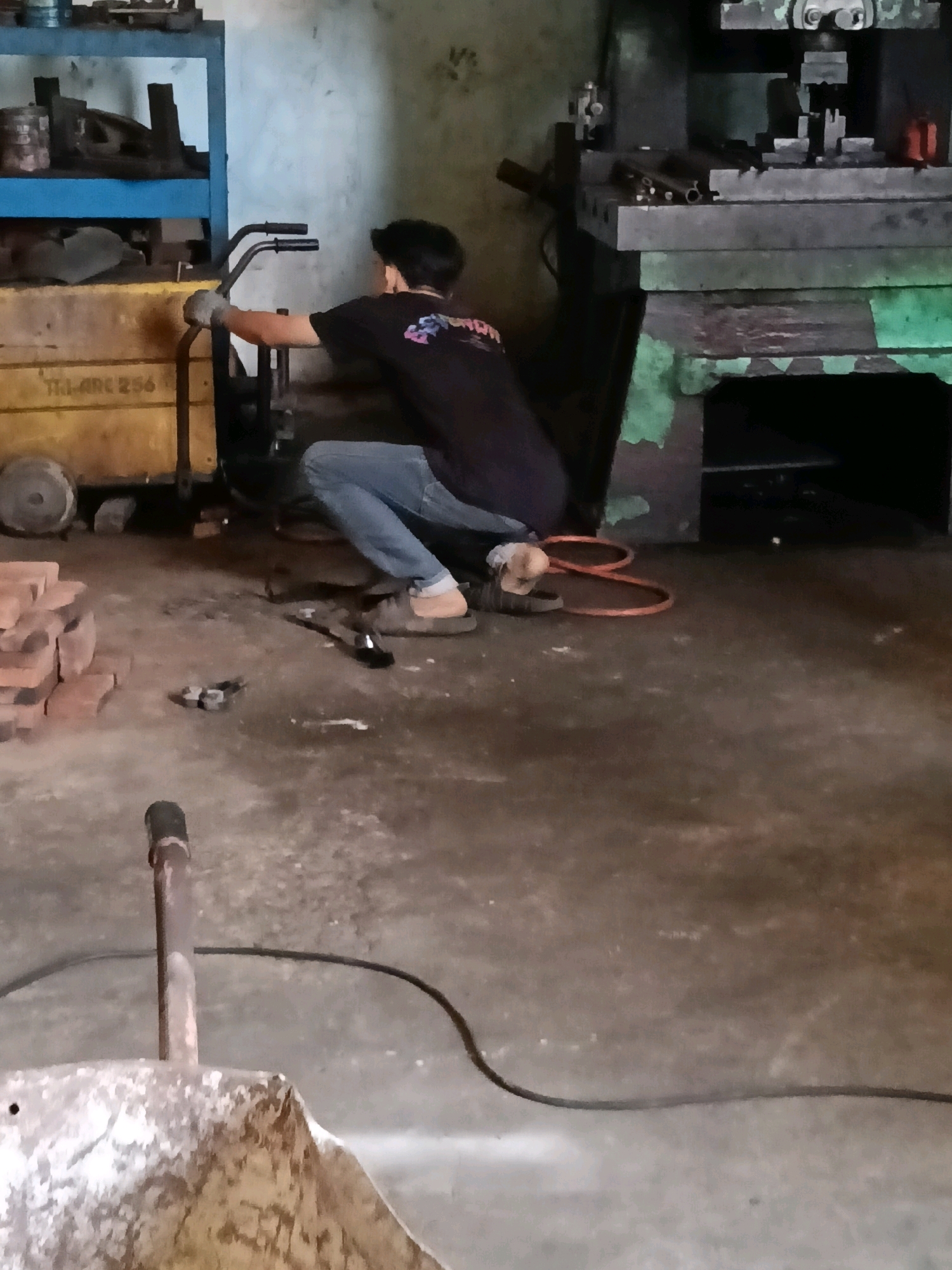 The image depicts an indoor scene, likely a workshop or factory, in Belimbing, Indonesia. A young man, wearing a dark t-shirt and light blue jeans, is crouched down and operating a piece of machinery with two black handles. He is wearing gloves on his hands. To his left, there is a yellow wheeled cart with "TL. ARC 256" visible on its side, and a blue shelf unit above it containing various metal objects. To his right, a large, dark green industrial machine with a metal platform is present. The floor is concrete and appears somewhat dirty, with scattered items including a stack of red bricks, tools, and a coiled orange cable. A black electrical cord snakes across the floor in the foreground. In the lower foreground, a section of a rusty metal implement, possibly part of a cart or shovel, is visible. The lighting appears to be artificial and somewhat dim.
