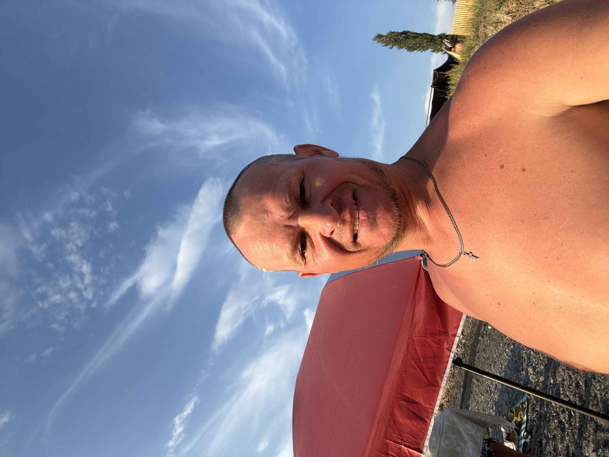 A shirtless man is taking a selfie outdoors, possibly at a beach or park. He has a shaved head, a light beard, and is wearing a silver chain with a cross pendant. Behind him, a large red umbrella is visible, along with a table covered with a white cloth. The background shows a blue sky with wispy clouds, a tall evergreen tree, and a fence. The ground appears to be gravelly. The lighting suggests it is daytime, possibly late afternoon given the warm tones on the man's skin. The location is identified as Zharbulak, Kazakhstan.