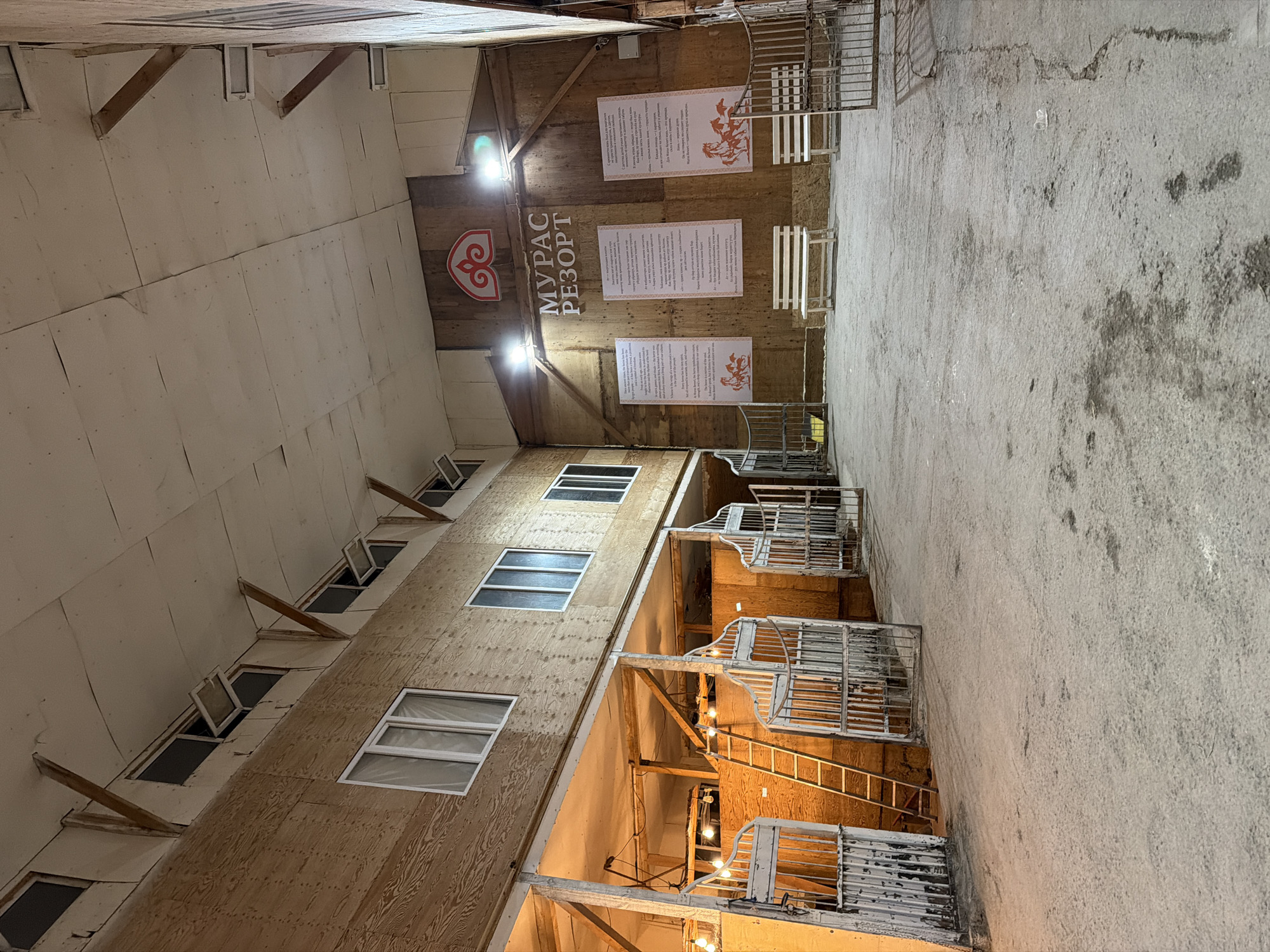 This media file depicts an interior view of a building, possibly an unfinished or rustic resort or stable, in Bishkek City, Kyrgyzstan. The scene is viewed from a low angle, looking upwards along a walkway or alleyway.
The subjects include the building's structure, characterized by wooden walls and partitions, and a concrete floor. Several metal cages or stalls with decorative tops are visible along the right side of the image, some with ladders leading to them. Small windows, some open, are set into the upper walls. A prominent logo with stylized red ornamentation and Cyrillic text reading "МУРАС РЕЗОРТ" is displayed on the wooden wall in the upper center, along with three framed posters containing text and illustrations. The lighting suggests it is either evening or an indoor setting illuminated by artificial lights, which cast a warm glow on the wooden surfaces and the cages. The overall impression is one of a functional, perhaps historical, or themed establishment.
There are no people visible in the image, nor is there any indication of weather. The location cues are limited to the Cyrillic text, which suggests a Cyrillic-speaking region, and the provided location context confirms it is Bishkek City, Kyrgyzstan. No specific emotions are conveyed by the static scene.