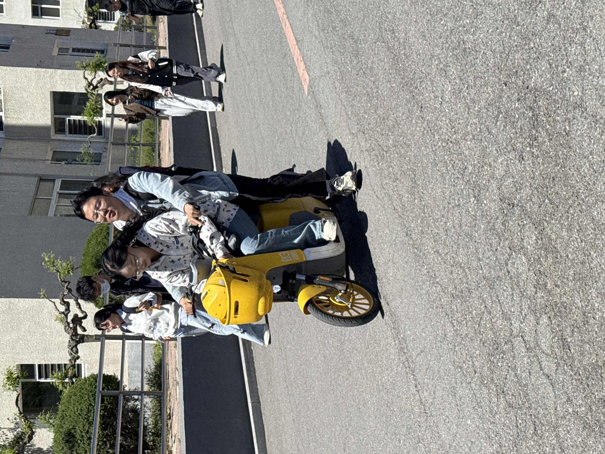 The image depicts a group of people riding a yellow scooter on a paved surface in Zhongtun, China. Two people are on the scooter, with additional people walking nearby. The location appears to be in an urban area, with buildings and landscaping visible in the background.