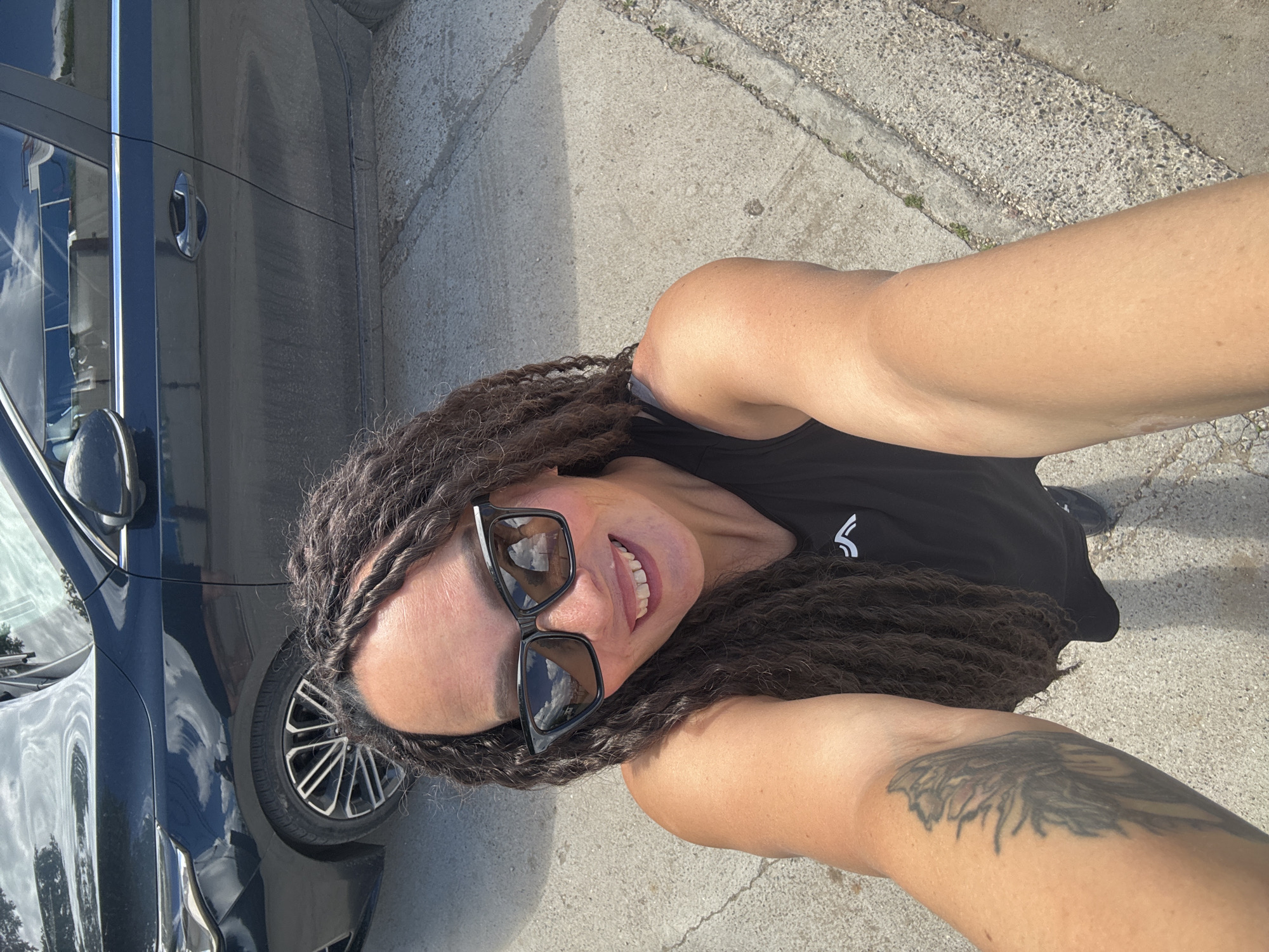 A medium, eye-level outdoor shot of a woman taking a selfie next to a dark blue car.
The woman is wearing a black tank top and sunglasses. She has dark hair in braids. She is smiling. A tattoo is visible on her right upper arm.
The car is parked on a concrete surface with a curb visible on the right side of the frame.
The location is outdoors on a sunny day. There is no information given within the prompt about this image being in Chaikovsky, Russia.