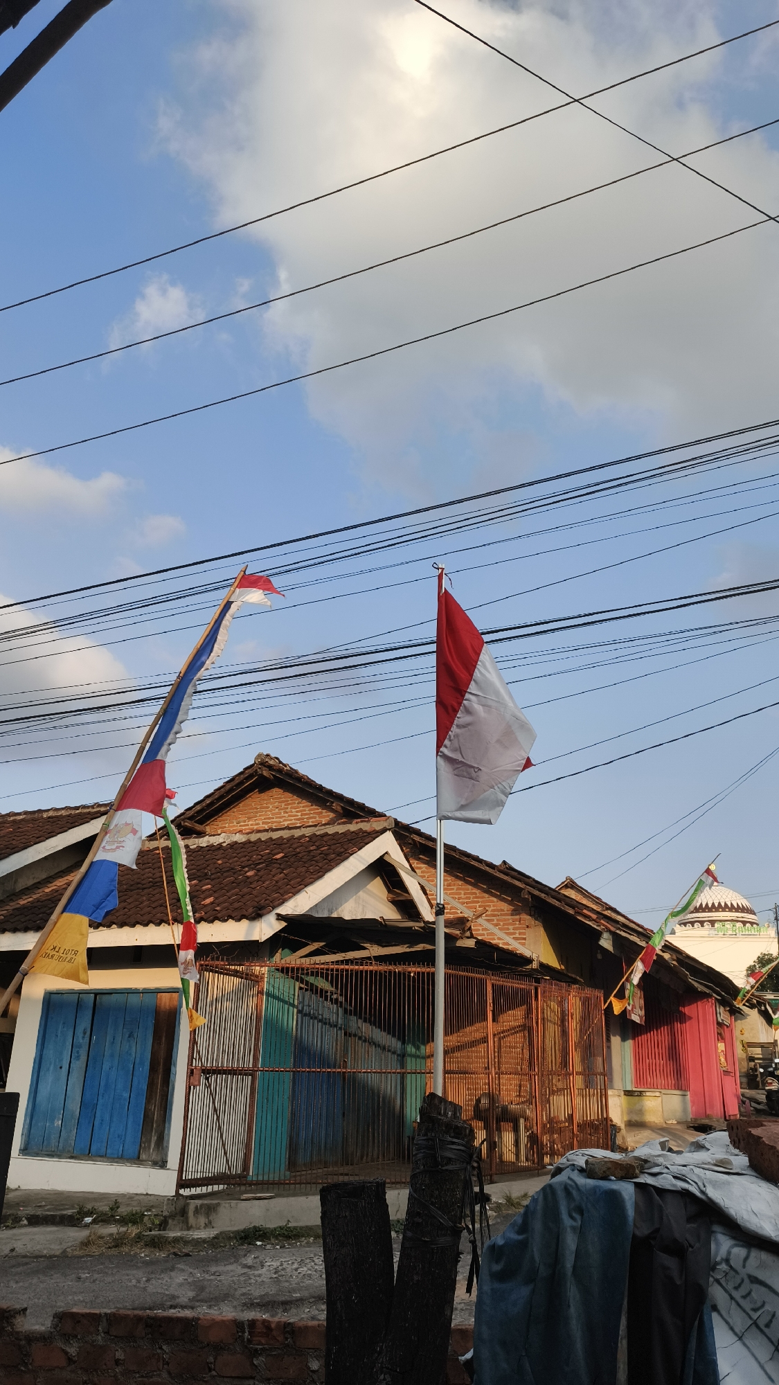 This is a photograph of a street scene in Bandar Lampung, Indonesia, featuring a row of buildings under a partly cloudy sky. The foreground includes a brick wall, some wooden posts, and tarpaulins covering debris. Behind this, a set of metal gates fronts a building with blue and teal doors. Further to the right, a building is painted bright red. Overhead, several power lines are strung across the sky. Two Indonesian flags are prominently displayed; one is a large red and white flag on a pole to the right, and another, a longer, multi-colored flag including red and white sections, is attached to a tall bamboo pole on the left. These flags suggest a celebration or national event. A domed structure, possibly a mosque, is visible in the background to the right, adorned with flags as well. The overall impression is a residential or commercial area in a Southeast Asian city, possibly decorated for a holiday.
