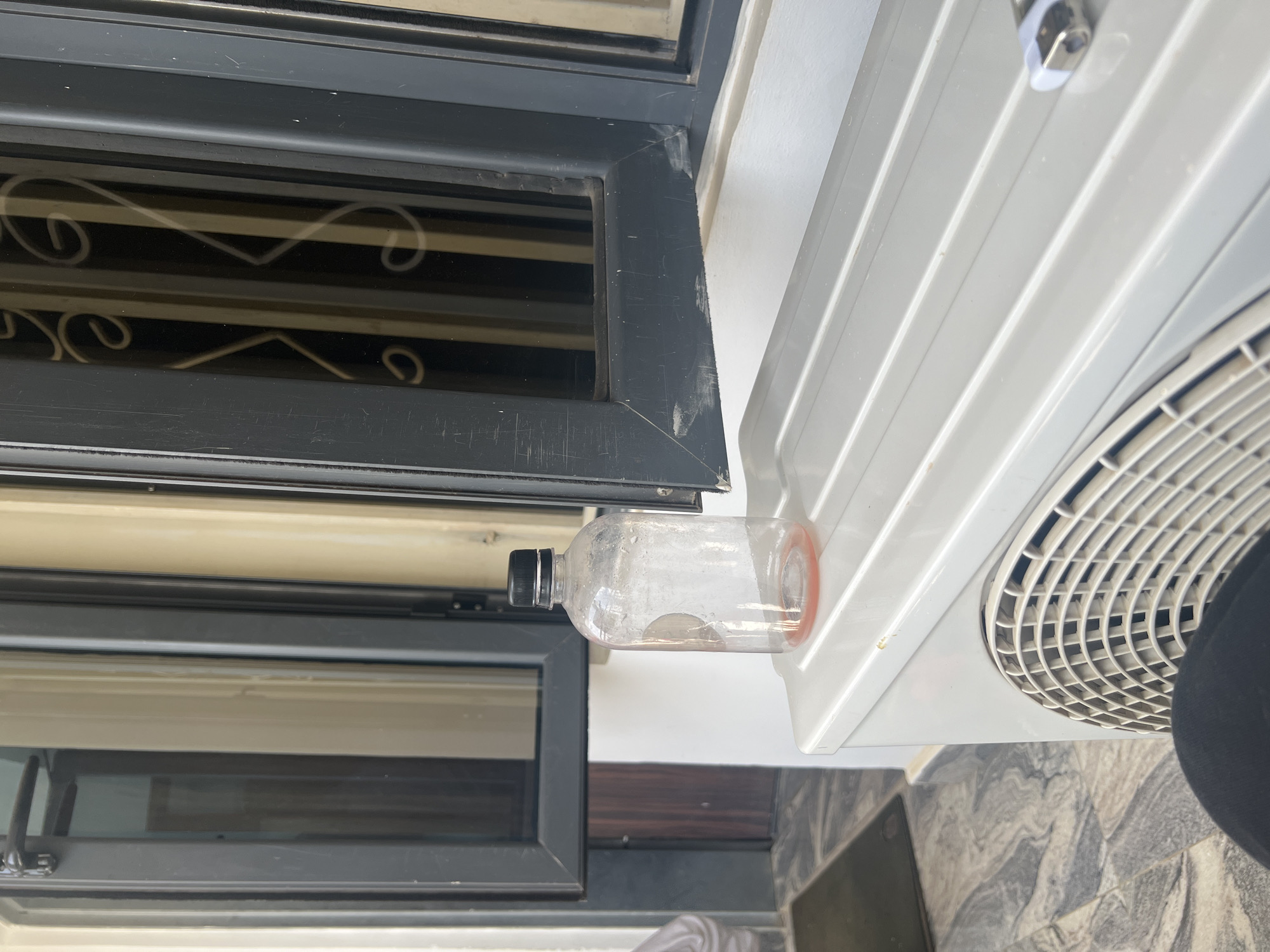 The image shows a close-up of a window frame, an air conditioning unit, and a plastic water bottle. The window frame is dark grey with decorative metal bars. The air conditioning unit is white, with a visible fan grill. A transparent plastic bottle with a black cap and an orange band is wedged between the window frame and the air conditioning unit. The background shows a wall, what appears to be a balcony floor and another window. The photo was taken in Gwarinpa, Nigeria.