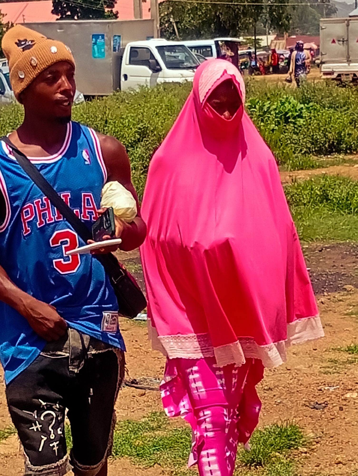 Two individuals are walking outdoors in what appears to be a casual, possibly rural setting. The person on the left is a young man wearing a tan beanie adorned with several shiny studs, a blue basketball jersey with "PHILA" and the number "3" visible, and dark denim shorts with white markings. He has a crossbody bag slung over his shoulder and is holding a phone and a white object in his hand. His expression is neutral, and he seems to be looking off to the side.
The person on the right is a woman wearing a vibrant pink head covering and a matching pink dress or robe with white lace trim along the hem. The garment has a flowing silhouette. The lower portion of her attire appears to be pink pants with a tie-dye or patterned design.
The background consists of a dirt path, green vegetation, and some vehicles in the distance, including a white truck and what looks like a minivan. There are also structures that suggest a marketplace or a populated area. The sunlight indicates it is daytime, and the presence of greenery suggests a warm climate. The overall atmosphere is that of a typical day in a community.