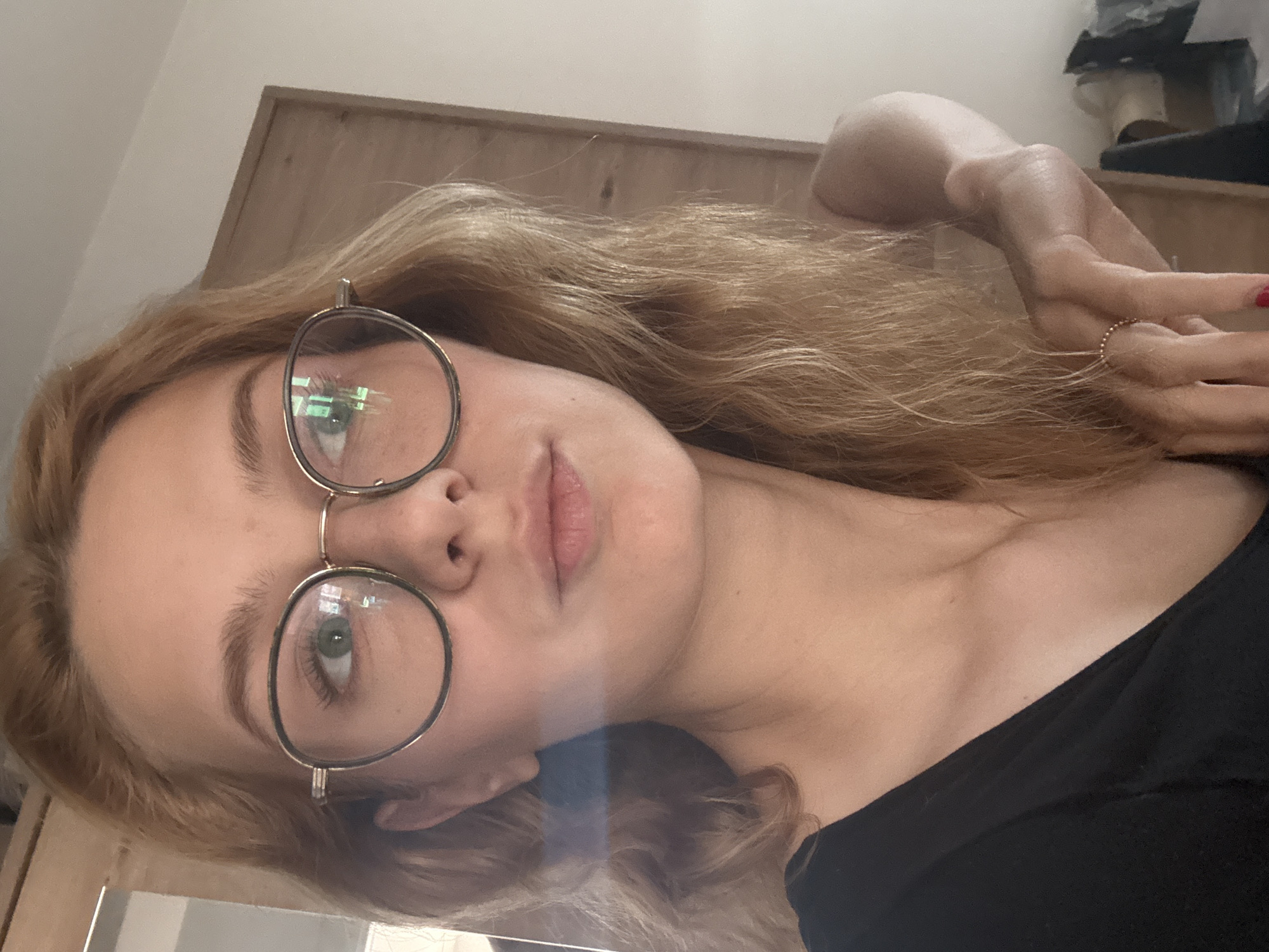 A young woman with blonde hair and glasses is looking into the camera. She is wearing a black top. She has a ring on her finger. The background appears to be indoors, with a wooden cabinet visible. The image does not contain information about the location in Kragujevac, Serbia.