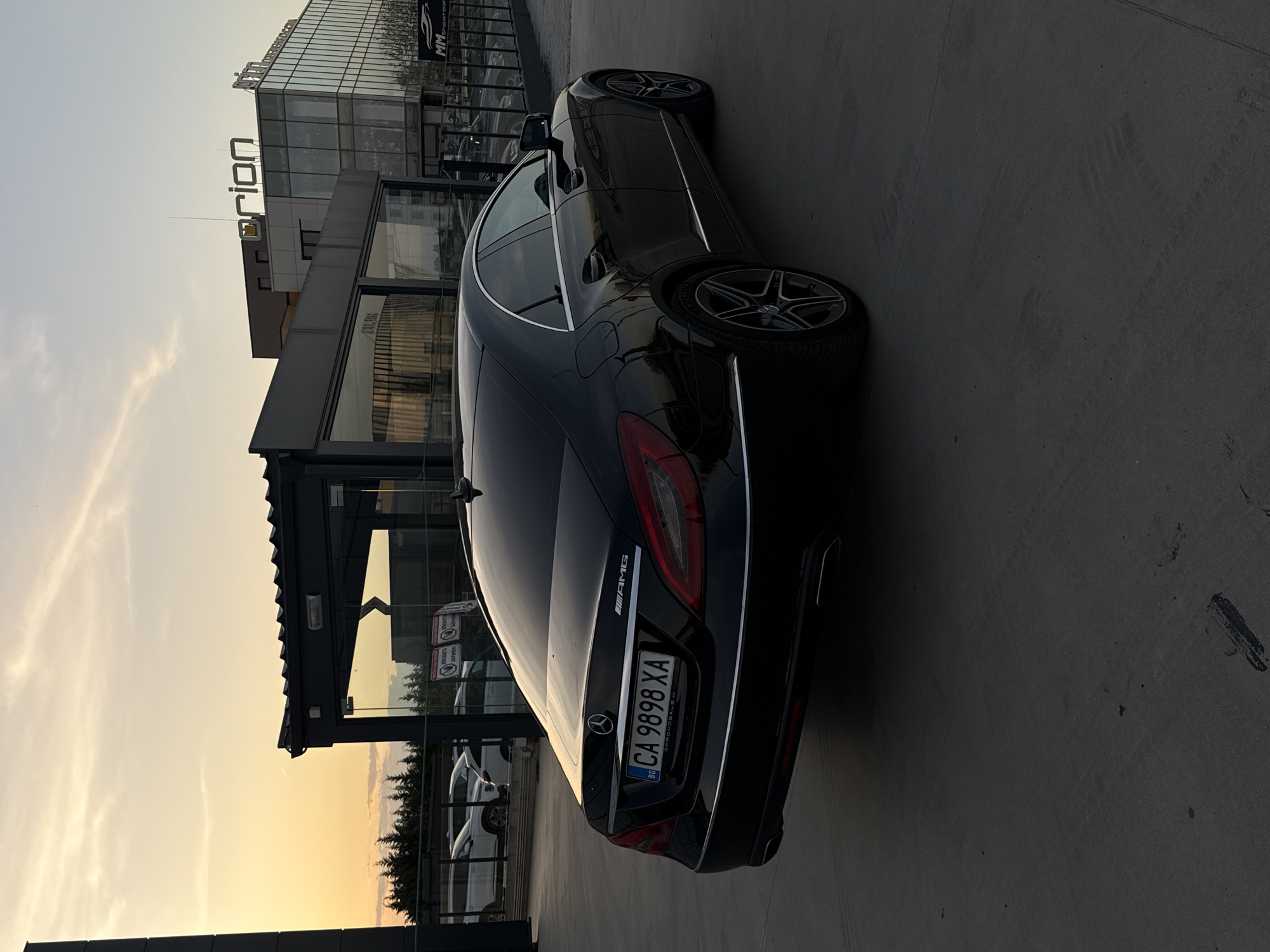 The image depicts a black Mercedes-Benz AMG sedan parked in front of the "Orion" building in Plovdiv, Bulgaria. The car's license plate reads "CA 9898 XA." The building behind it appears to be a modern structure, possibly a car dealership or showroom, as indicated by the "MM Showroom" sign. The sky is a mix of light colors, suggesting either sunrise or sunset.