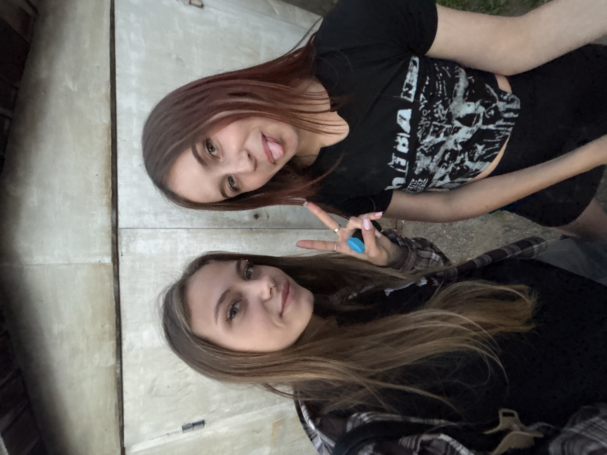 Two young women are posing for a photograph in what appears to be an urban setting. The woman on the top has brown hair with reddish highlights and is wearing a black t-shirt with a white graphic design. She is sticking her tongue out and holding up a peace sign. The woman on the bottom has long blonde hair and is wearing a black top with a plaid shirt. She has a nose piercing and an eyebrow piercing. They are standing against a weathered concrete wall. Based on the provided location, the picture was taken in Фоки, Russia.