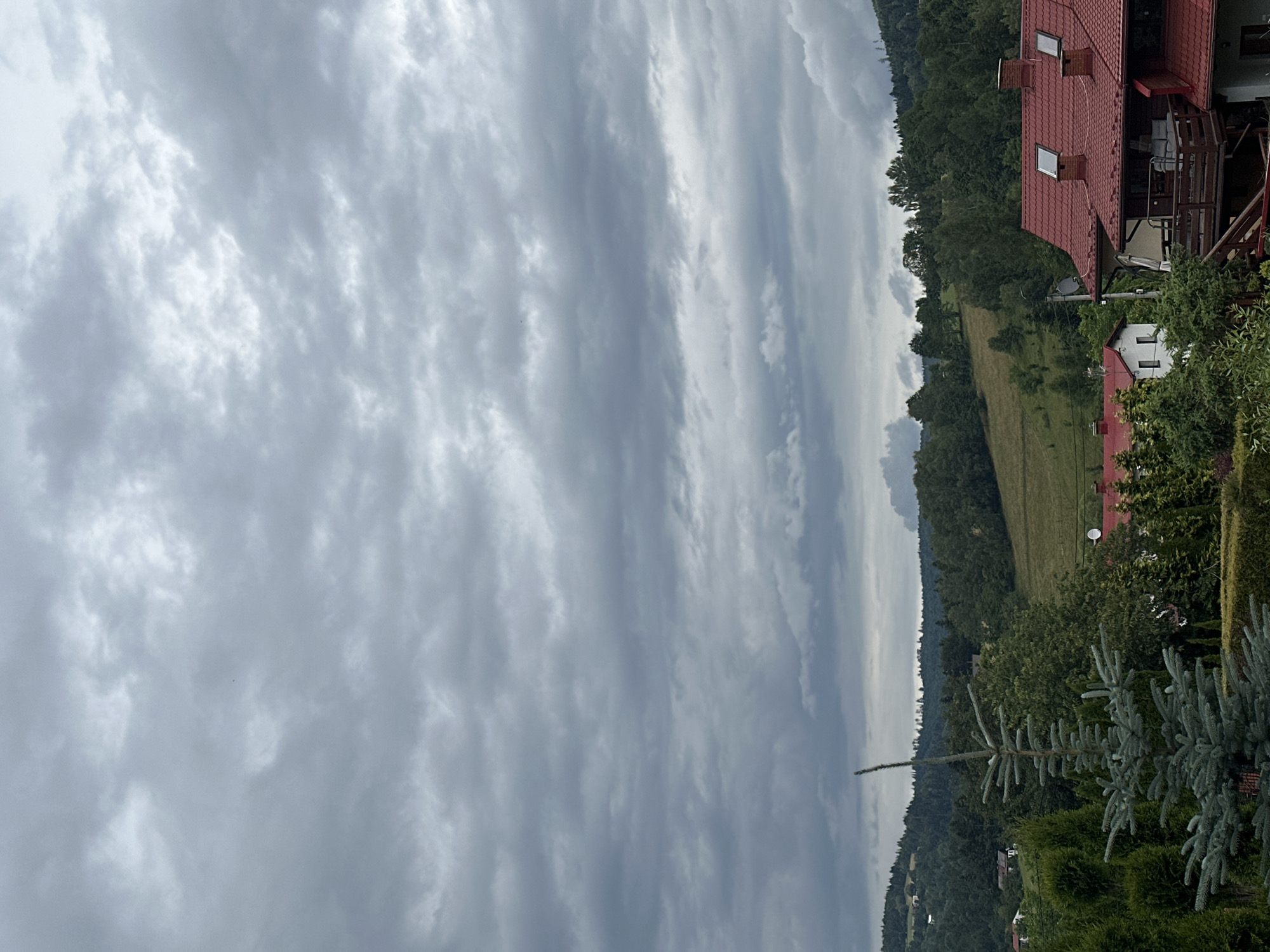 This is a photograph taken in Wisła, Poland, on an overcast day. The image is oriented vertically, with the sky occupying the majority of the frame and a landscape with houses and trees filling the lower portion.
The sky is filled with a dense layer of grey clouds, suggesting a cloudy or possibly rainy day. The clouds are textured, with some lighter patches indicating variations in thickness and light penetration.
The landscape below shows a hilly terrain covered in lush green trees. A few houses with red tiled roofs are visible nestled amongst the trees. One house in the foreground has a prominent red roof with dormer windows and a wooden balcony. Another smaller white house with a red roof is also visible. The vegetation appears dense and verdant, characteristic of a rural or semi-rural setting. The presence of trees and the rolling hills indicates a natural, possibly mountainous or hilly region.
There are no people visible in the image. The primary focus is on the natural scenery and the residential structures. The lighting is diffused due to the overcast sky, casting a soft, even light on the landscape. The time of day is not explicitly indicated but appears to be daytime. There is no visible text in the image.