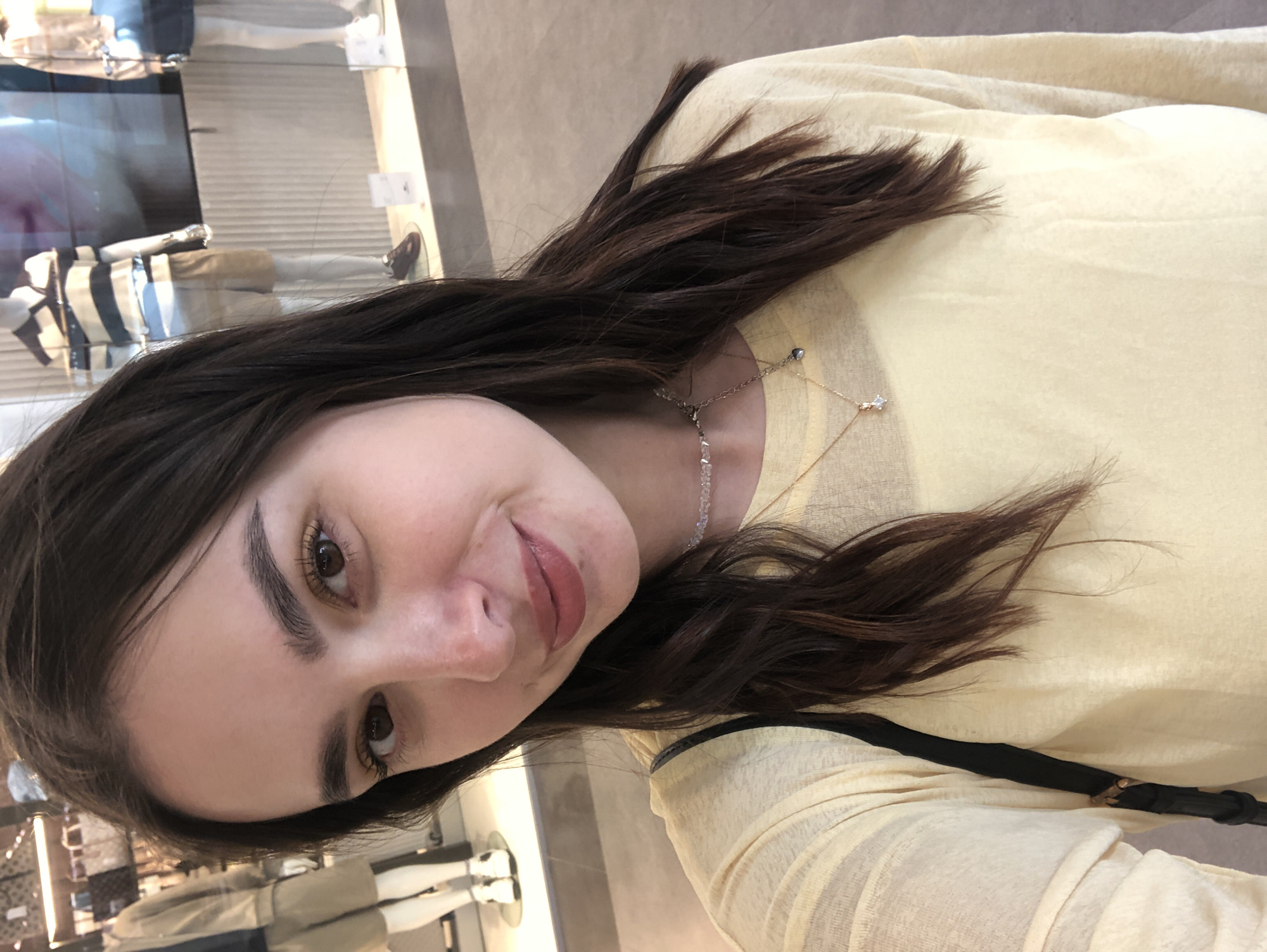 This media file is a close-up, rotated selfie of a young woman with long, dark brown hair. She is wearing a light yellow, long-sleeved top and a delicate necklace. Her expression is a soft smile, and she appears to be looking slightly to her left. The background is blurred, but it suggests an indoor setting, possibly a store or mall, with mannequins and merchandise visible. There is no visible text in the image. The lighting is soft, indicating an indoor environment during daylight hours. The overall impression is casual and personal.