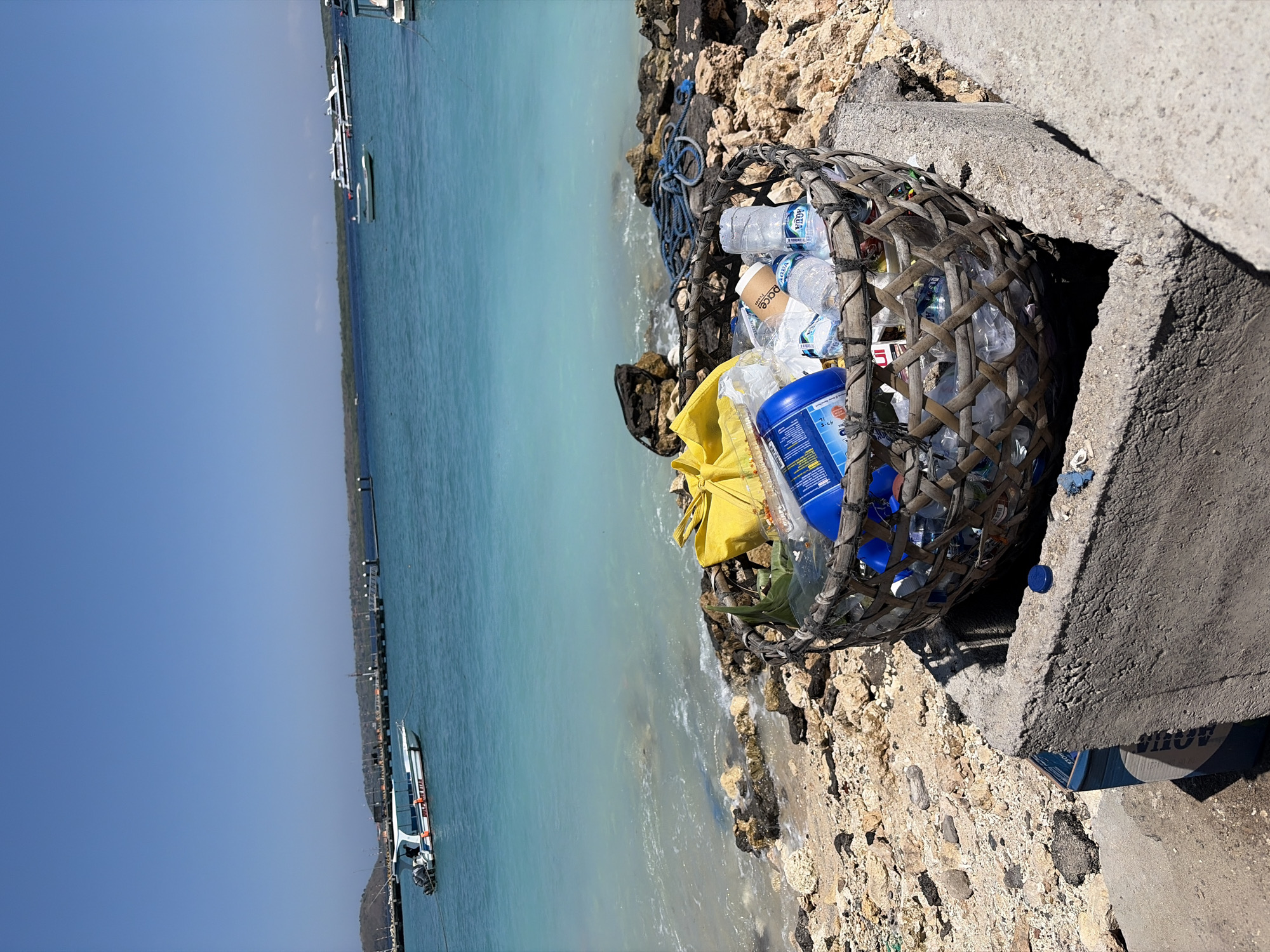 A woven basket overflows with plastic bottles, bags, and other refuse, perched on the edge of a concrete structure near a rocky shoreline. The turquoise water of the ocean stretches out beyond, dotted with boats and a distant pier. The sky above is a clear, vibrant blue, suggesting a sunny day. The scene, located in Nusa Penida, Indonesia, highlights the issue of plastic pollution on the island.