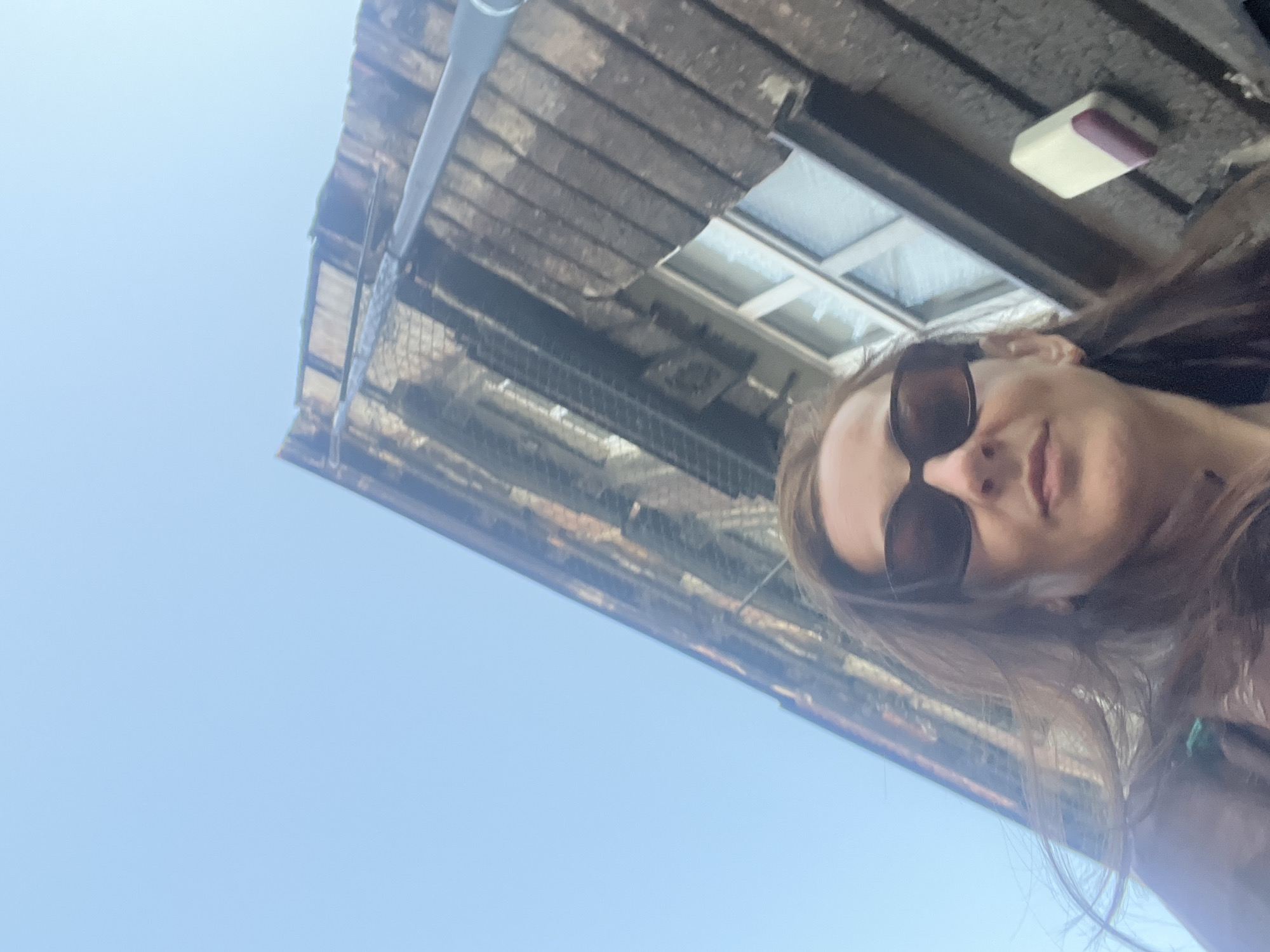 A low-angle, outdoor, wide shot shows a woman from the chest up in the foreground, looking directly at the camera with sunglasses on. The woman has long, brown hair and is wearing a dark top. Behind the woman, an old, weathered building with dark siding and a white-paned window fills the upper portion of the frame, angled upwards toward a clear blue sky. The building appears to have a fire escape or railing running vertically up its side.