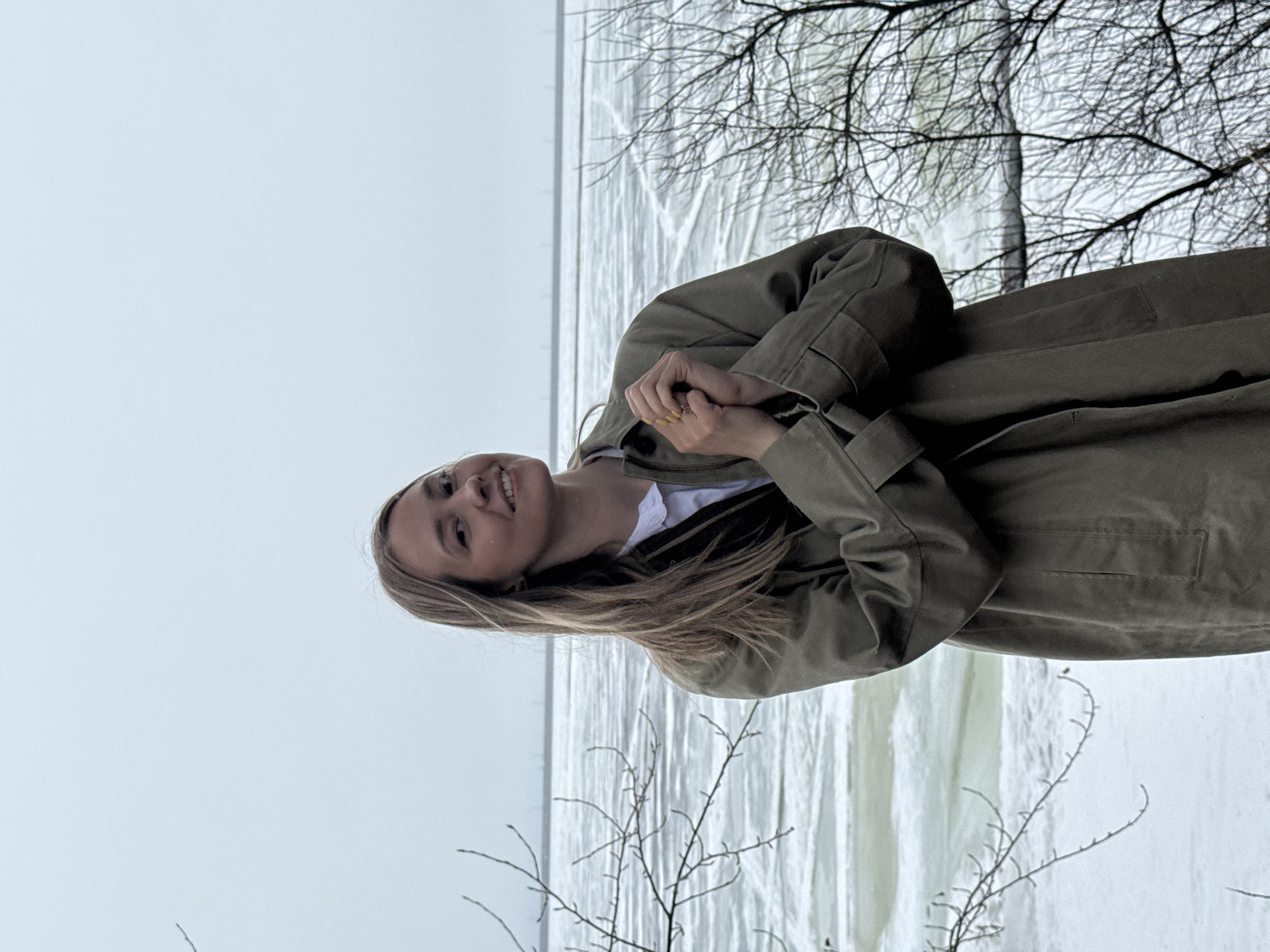 The image features a young woman standing outside, possibly in a park or natural area. She is wearing a light olive-green trench coat over a white shirt. Her blonde hair falls loosely around her shoulders. Her hands are clasped together in front of her. She is smiling.
Behind her, there is a partially frozen body of water, which appears to be a river or lake, with ice fragments on the surface. In the foreground, thin, bare branches of trees or bushes are visible. The sky is overcast and grey, suggesting a cold or winter day.
Based on the metadata, the image was taken in the city of Chaikovsky, Russia.