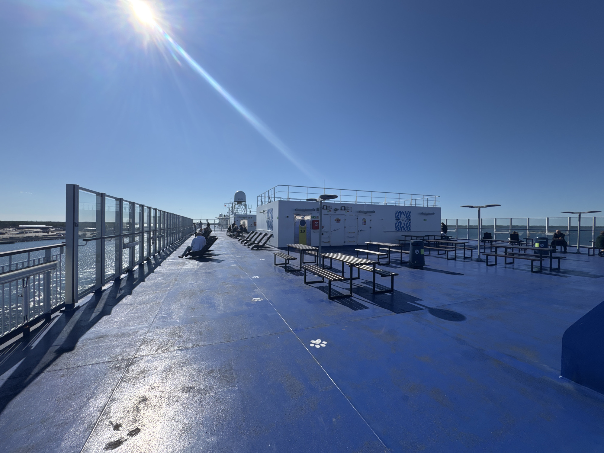 The image depicts the open deck of a ferry, likely docked or at sea near Karlskrona, Sweden, under a clear blue sky. Sunlight streams in from the upper left, casting long shadows across the bright blue deck. Several picnic tables with attached benches are scattered across the deck, some empty and others occupied by a few individuals.
To the left, a railing and a glass barrier separate the deck from the calm water, where a hint of land and structures can be seen in the distance, suggesting proximity to shore or a harbor. The ferry's white superstructure, featuring communication equipment like radar dishes, is visible in the background. White paw prints are painted on the deck at regular intervals.
On the deck, a few people are seated on benches or lounge chairs, some looking out at the water, others engaged in conversation or reading. The scene is orderly and serene, characteristic of a ferry crossing on a sunny day. The presence of the paw prints might indicate a designated area for pets or a playful design element.