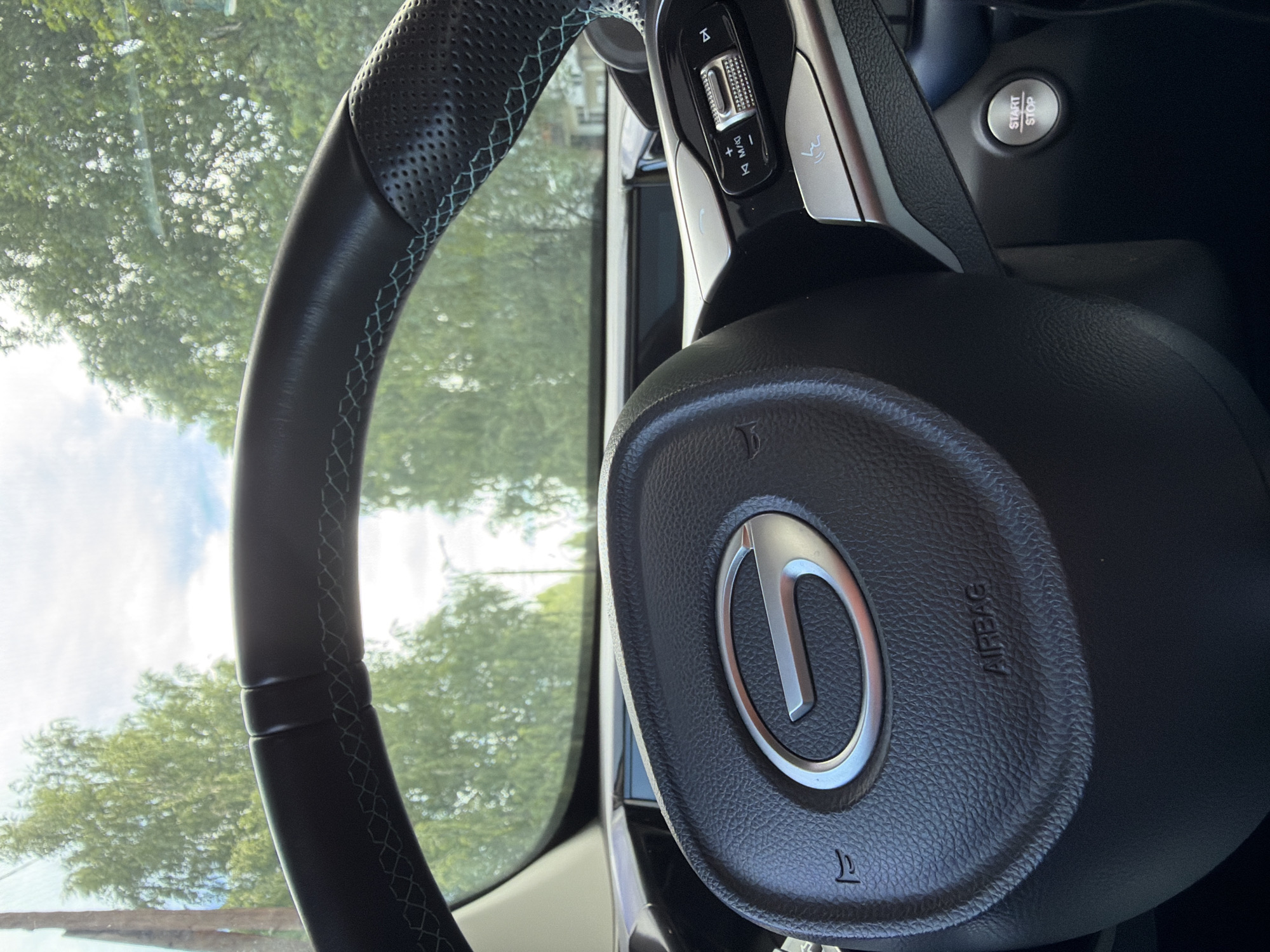 The image shows a close-up view of a car's steering wheel, viewed from the driver's seat. The steering wheel is black leather with light blue stitching along the top edge. The car's logo, a stylized 'G' in a silver oval, is prominently displayed in the center of the steering wheel. The word "AIRBAG" is visible on the lower part of the steering wheel. To the right of the steering wheel are various controls, including buttons and a dial, suggesting the presence of infotainment and cruise control functions. Below these controls, a "START/STOP" button is visible. The background of the image, seen through the car's windshield, shows a bright, slightly overcast sky with green trees, suggesting an outdoor environment. There are no people visible in the image. The scene appears to be a parked car, ready for operation. The location in the city of Chaikovsky, Russia is not determinable from the visual information provided in the image.
