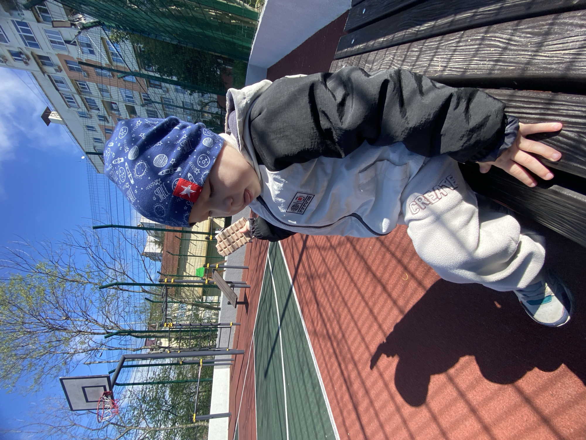 A child wearing a blue hat and a jacket is sitting on a wooden bench. The child is near a basketball hoop and a fenced area, indicating a playground or recreational area. The ground is marked with red and green lines, possibly a sports surface. In the background, there is a residential building, which suggests the location is in a city. The sky is blue with some clouds, indicating it's daytime. The presence of trees, some with budding leaves, suggests it might be springtime. Based on the provided information, it is not possible to confirm that the location is Sochi, Russia.