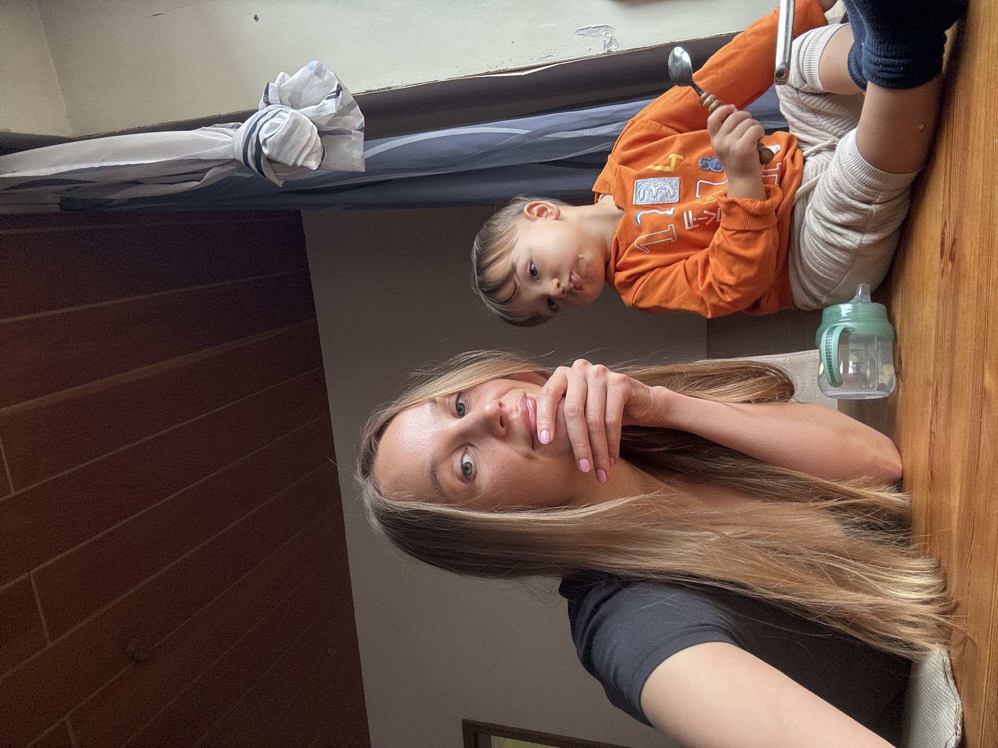 A blonde woman with long hair and pink fingernails is seen in a close-up selfie, resting her chin on her hand. Beside her, a young boy with brown hair is wearing an orange hoodie with white and blue numbers and beige pants. He is holding a spoon and looking off to the side. They are sitting at a wooden table, with a window and curtains visible in the background. A baby bottle is also on the table. The location appears to be a domestic setting within the city of Überherrn, Germany.