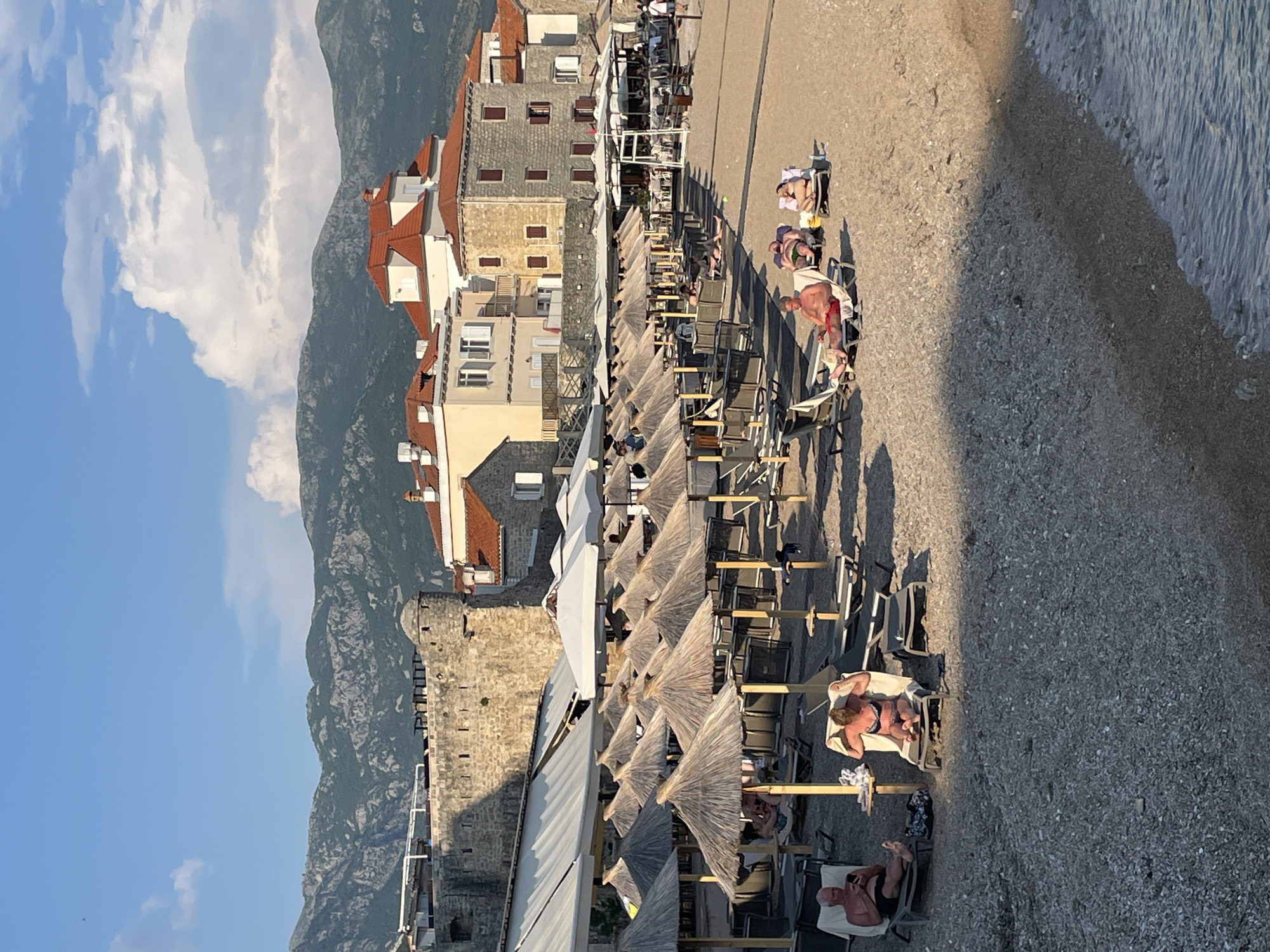 The image depicts a beach scene in Budva, Montenegro, with mountains visible in the background under a partly cloudy sky. The beach is lined with rows of sunbeds and thatched umbrellas. People are relaxing on the sunbeds, some sunbathing and others sitting. Adjacent to the beach is a series of buildings with red-tiled roofs and stone facades. A stone wall or fortification also runs parallel to the beach. The sea is visible on the right side of the image.