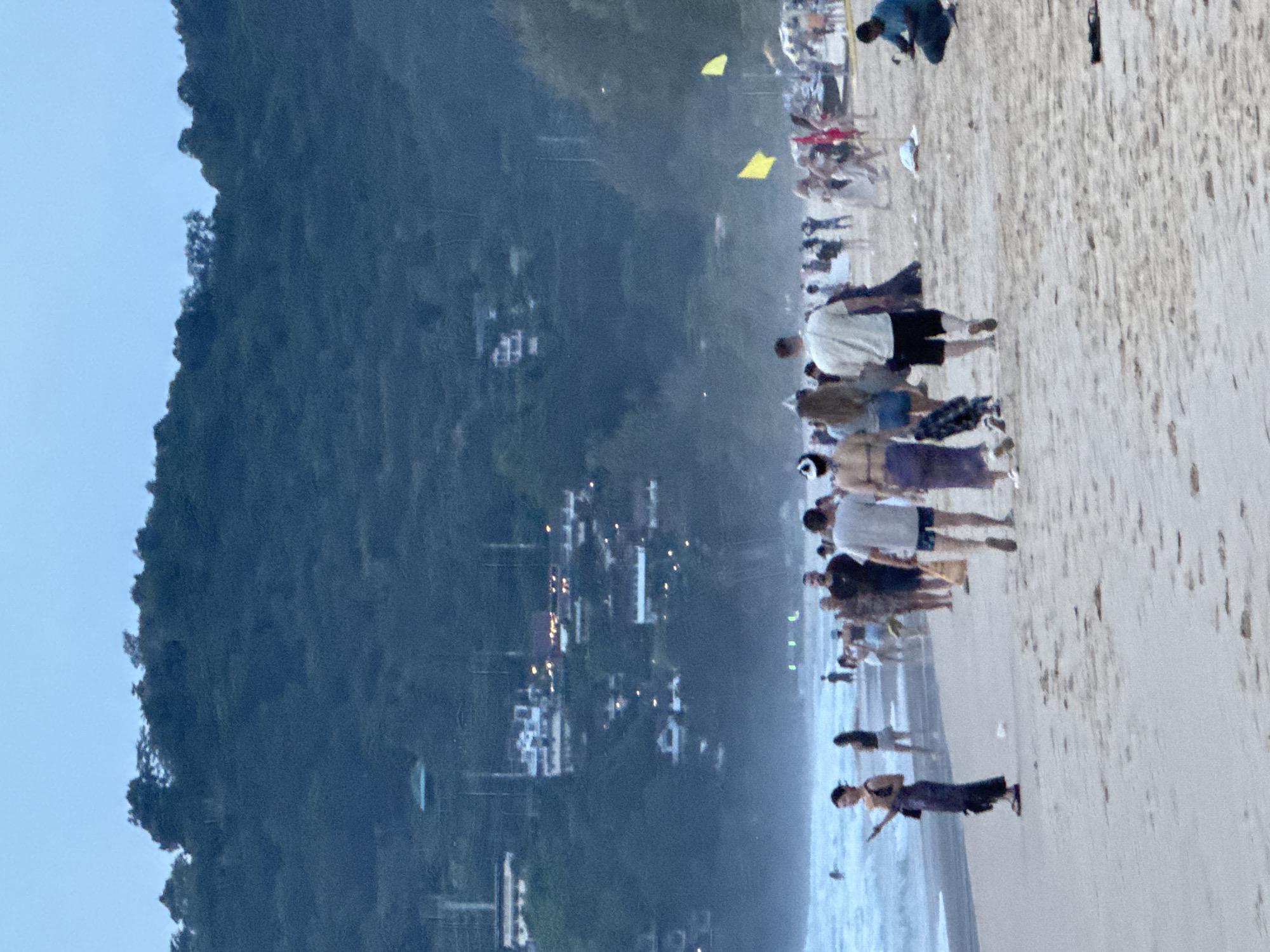 A group of people are walking on a sandy beach in Karon, Thailand. The sky is overcast and it appears to be dusk. The ocean is visible in the background, with waves rolling onto the shore. There are buildings on a hill in the distance. Some of the people are wearing swimsuits, while others are wearing casual clothing. Some are walking alone, while others are in groups. There are a few yellow flags and red flags on the beach.