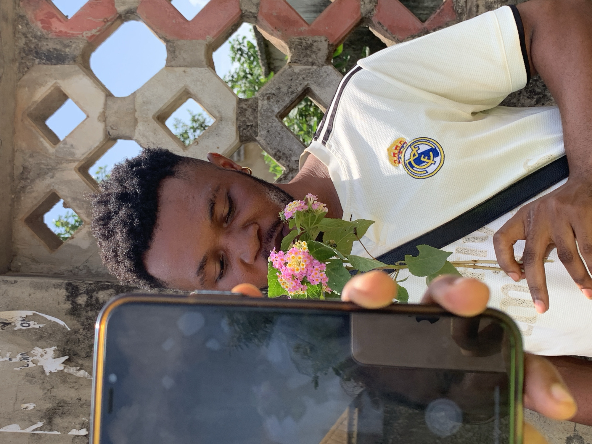 A medium shot depicts a dark-skinned man holding a smartphone and a pink and yellow flower. He is wearing a white t-shirt with black stripes on the shoulders and a Real Madrid club logo. The scene is set against a weathered concrete wall with red and grey geometric patterns. The background shows hints of green vegetation and a clear blue sky. The image is taken outdoors in daylight. Although the prompt mentions Ikorodu, Nigeria, there is no information in the image itself to confirm that location.
