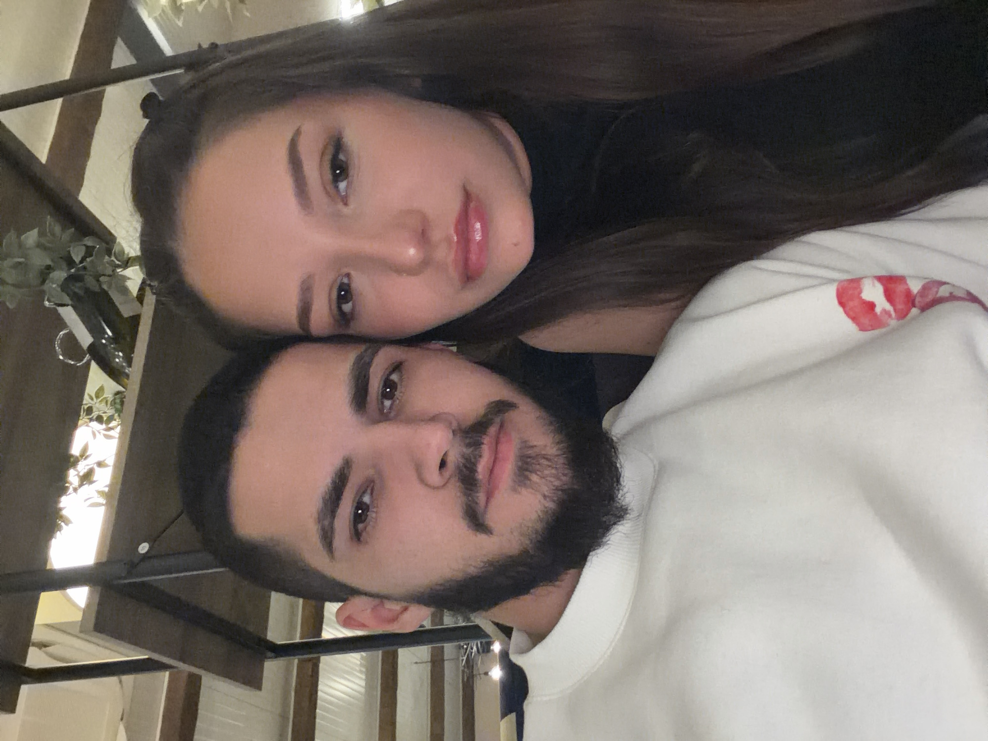 The image displays a posed, close-up photograph of two individuals, an adult male and an adult female, situated indoors within an establishment in Varna, Bulgaria. The male, positioned on the left, has dark hair, a trimmed dark beard and mustache, and is wearing a light-colored crew-neck sweatshirt featuring a red lip print graphic on its left sleeve. The female, positioned on the right, has long dark brown hair and is wearing a dark, likely black, sleeveless top. Both individuals are looking directly at the camera. The background consists of indistinct indoor elements, including a dark-framed shelving unit with a visible wooden shelf, and artificial green foliage used as decorative elements. The lighting indicates an indoor setting.