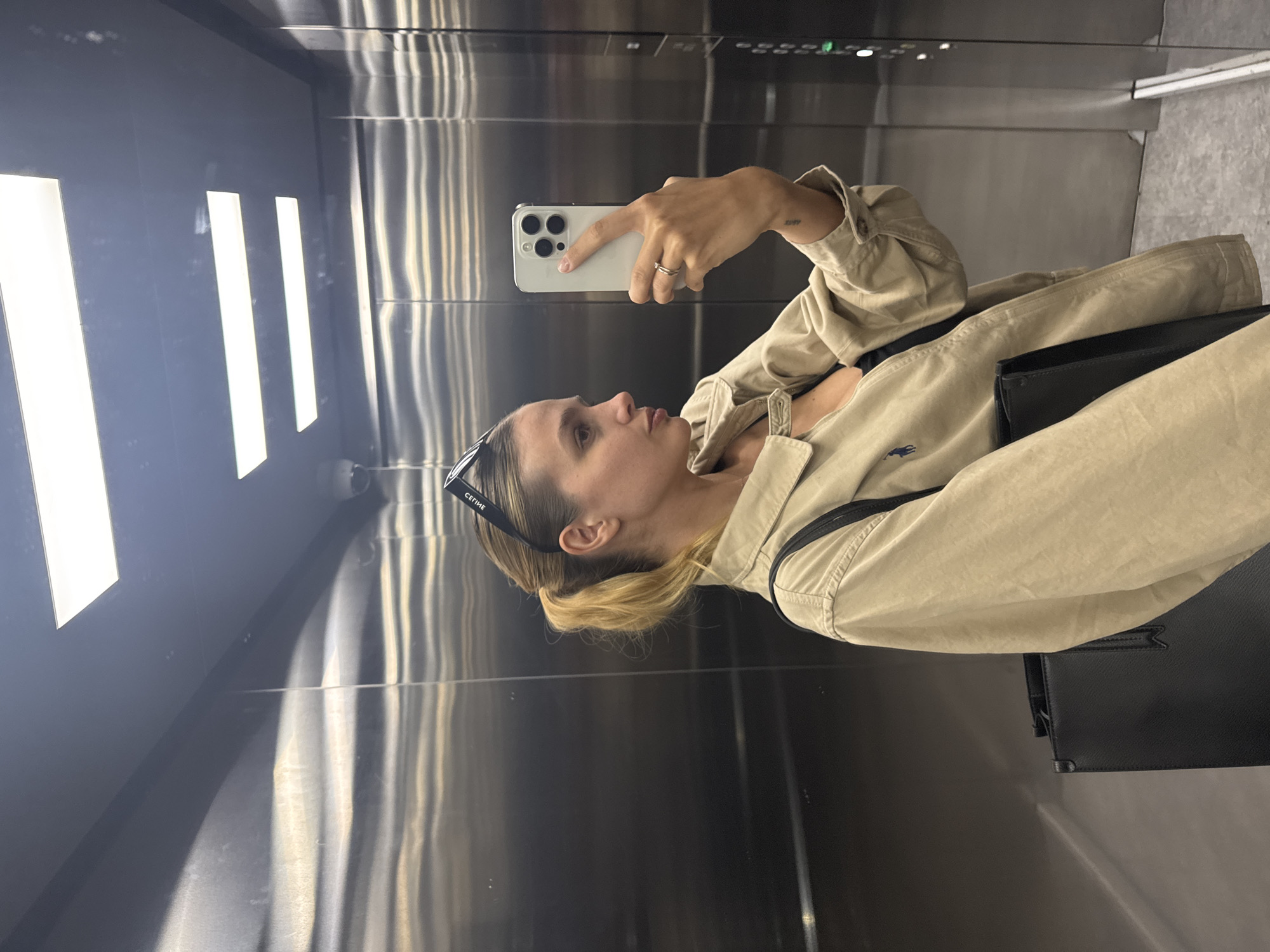 A blonde woman wearing sunglasses on her head is in an elevator in Unknown, Unknown. She's holding a smartphone, presumably taking a selfie. She's wearing a beige collared shirt and has a black purse slung over her shoulder. The elevator has metallic walls with vertical striations and built-in rectangular light panels.