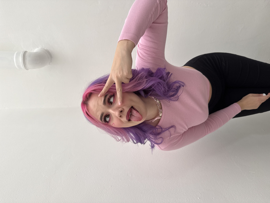 The image depicts a female individual, presumed to be in the City of Zagreb, Croatia, posing against a stark white interior wall. She has shoulder-length, wavy hair dyed vibrant pink on top and purple transitioning to lighter purple/lavender on the ends. Her eyes are crossed, looking upwards and to the right, and her tongue is extended. A small pink heart is drawn on her left cheekbone. She is wearing a light pink, long-sleeved cropped top and black pants. A necklace made of small, light-colored shells or irregular beads is visible around her neck. Her left hand forms a peace sign (V-shape) with the index and middle fingers, positioned near her left eye. The background consists of a plain white wall forming an interior corner, with a white pipe featuring an elbow joint visible high on the right side.