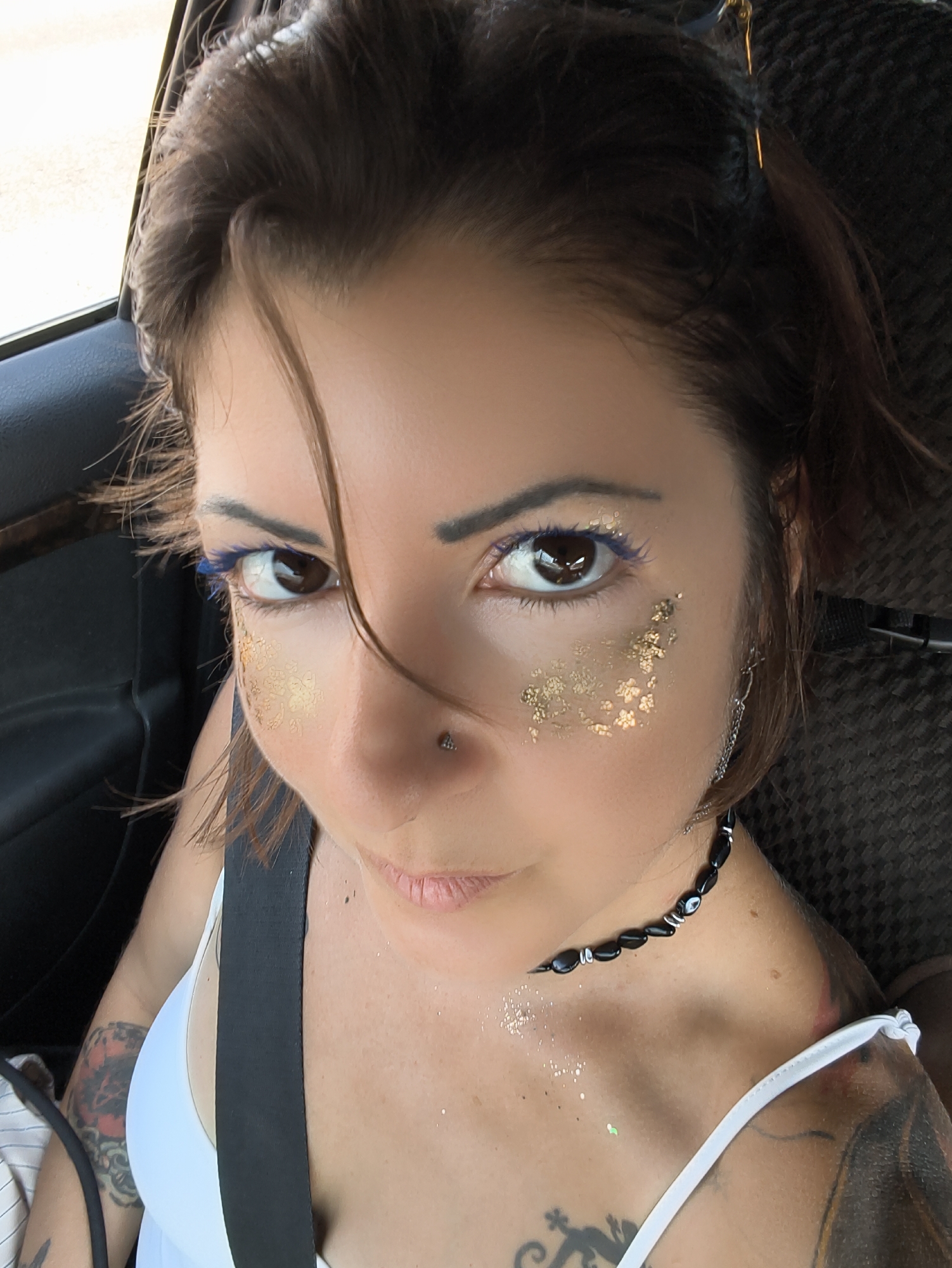 A young woman with brown hair and fair skin is shown in a close-up shot, looking directly at the camera. She is wearing a white top and a black beaded necklace. Her eyes are brown and she has blue eyeliner and gold glitter makeup on her cheeks. She has a nose piercing and several tattoos on her shoulders and chest. The background appears to be the interior of a car, with a dark-colored seat visible behind her. The setting is likely a public road in the Ramat Negev Regional Council, Israel, possibly during daylight hours given the lighting conditions.