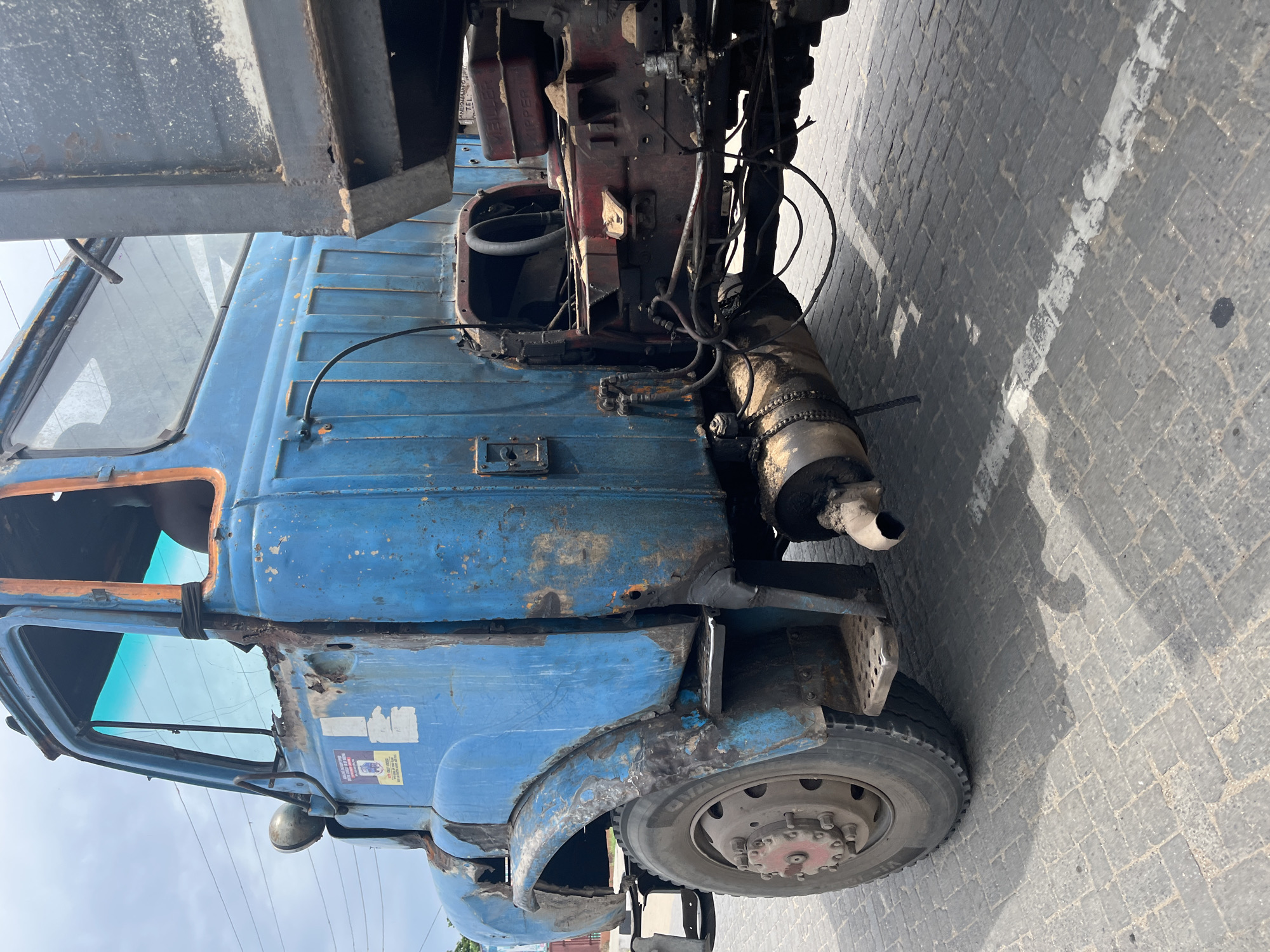 An aged and visibly rusting blue truck is positioned on a cobblestone street in Badagry, Nigeria. The truck's cab is heavily corroded, particularly around the wheel wells and door frames. The vehicle's engine is exposed. The truck appears inoperable. The setting is urban, and the lighting suggests it is daytime. No people are visible.