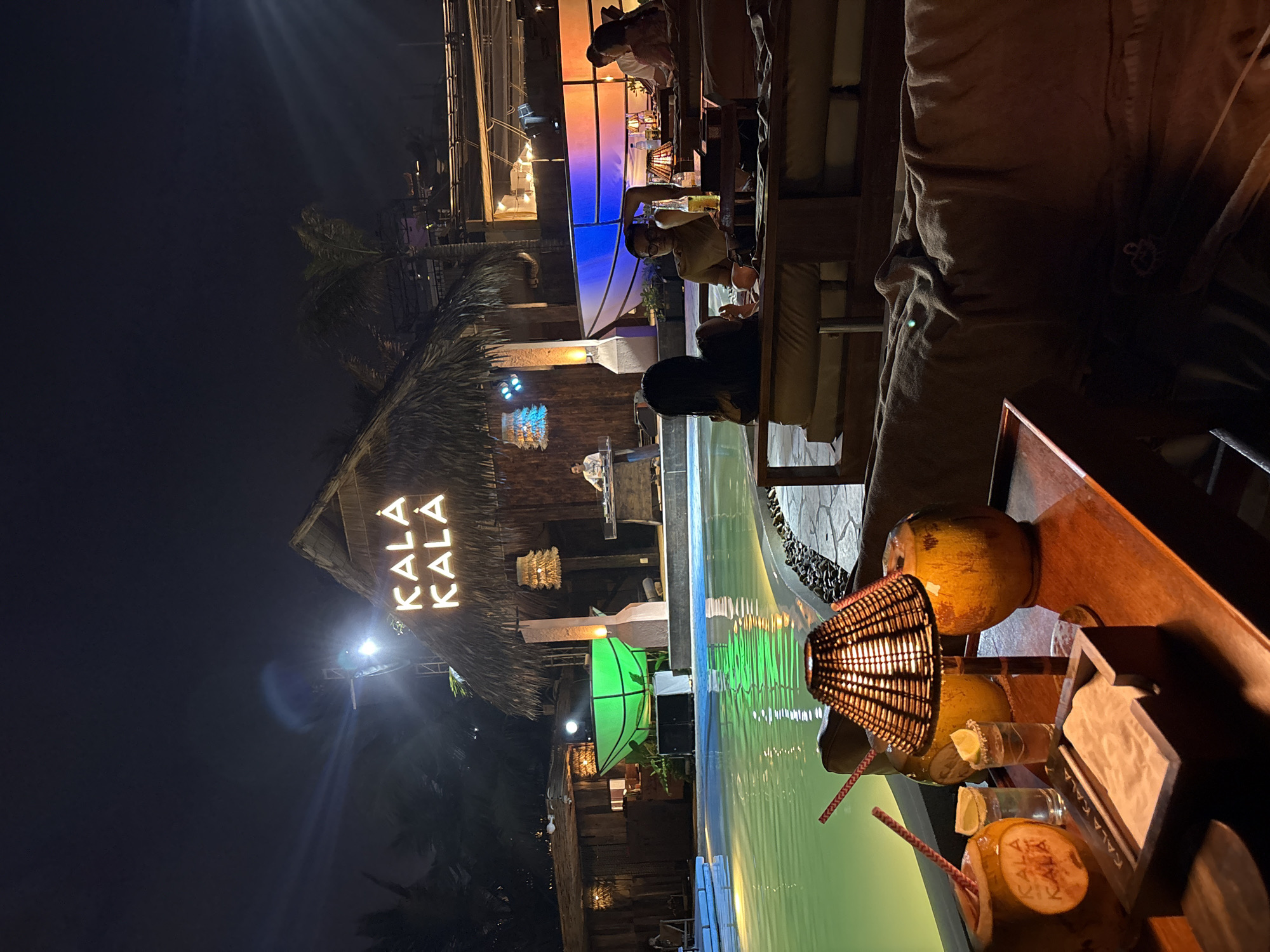 The image depicts an outdoor setting at night, likely in a resort or upscale dining venue. The foreground shows a wooden table with drinks served in coconuts, garnished with lime and straws. A woven lamp adds to the ambiance. Adjacent to the table is a pool that reflects green hues.
In the background, the thatched roof and illuminated "KALA KALA" sign indicate the name of the establishment. The scene is populated with people seated and standing, appearing to be patrons enjoying themselves. A DJ booth is visible, suggesting live entertainment.
Based on the name, "KALA KALA," and the environment depicted, the location is potentially in Đà Nẵng, Vietnam, known for its beach resorts and nightlife.
