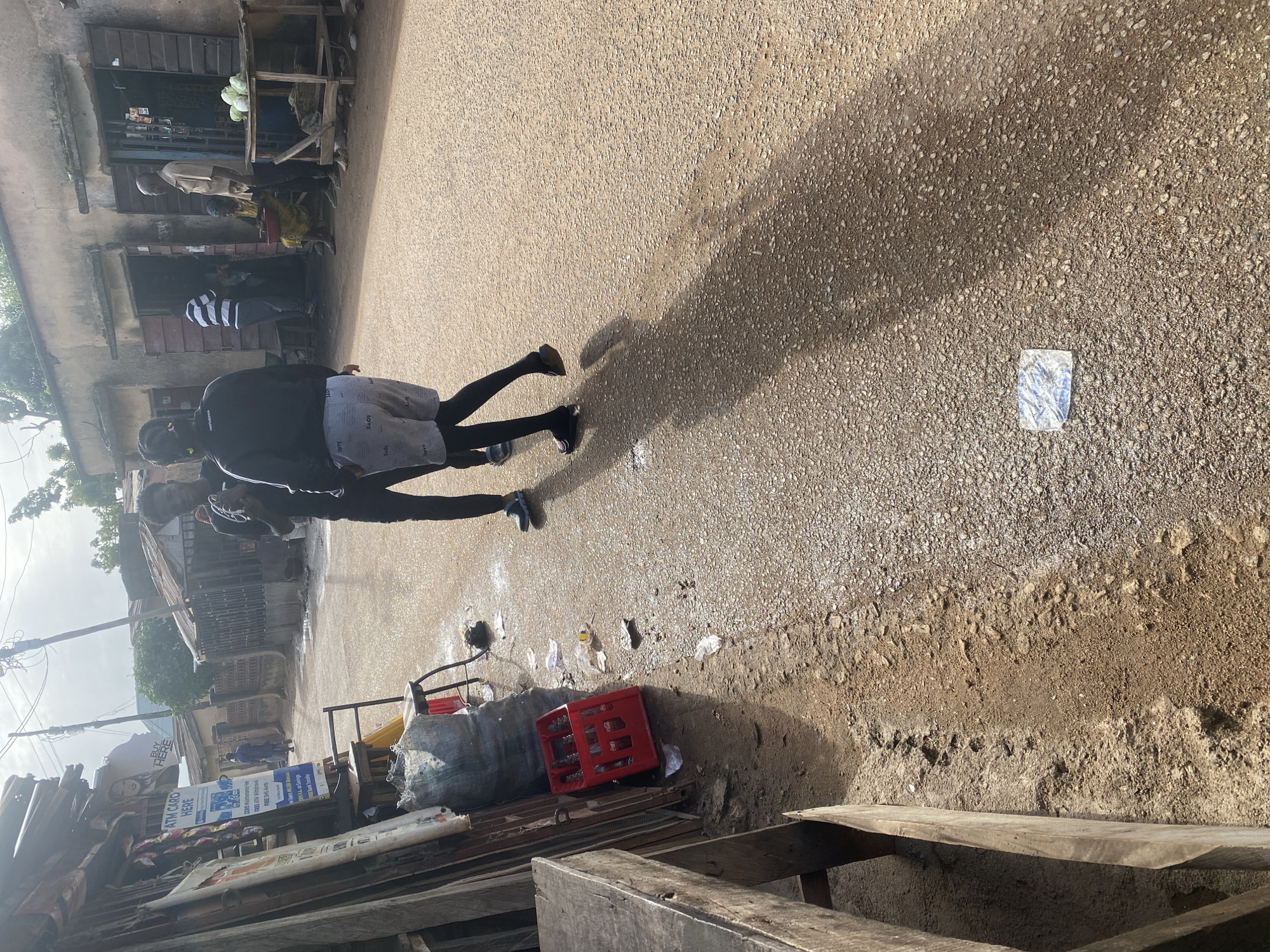The image depicts a street scene in Jos, Nigeria, likely during daylight hours given the strong shadows cast by the people and objects. The foreground is dominated by a rough, gravelly asphalt road, with patches of dirt and debris. To the left, a collection of wooden structures, possibly market stalls or shops, are visible, some with goods displayed. A large, grey, lumpy bag is prominent, along with a red plastic crate that appears to hold bottles.
In the mid-ground, two individuals are walking away from the camera, their bodies angled towards the shops on the left. One person is wearing a black hooded jacket and grey shorts, while the other is wearing a black jacket over a striped t-shirt and grey pants. They are casting long shadows on the road. Further down the street, other figures are less distinct, some near the shop fronts.
Several pieces of visible text offer clues to the location's context. A sign reads "ATM CARD HERE" with additional text below about maintenance fees, withdrawals, and savings, suggesting a commercial area where financial services are offered. Another sign with "BUY HERE" also indicates a retail setting. The overall atmosphere appears to be that of a busy, possibly informal, marketplace or commercial street. The weather seems clear, with the sun high enough to cast pronounced shadows.