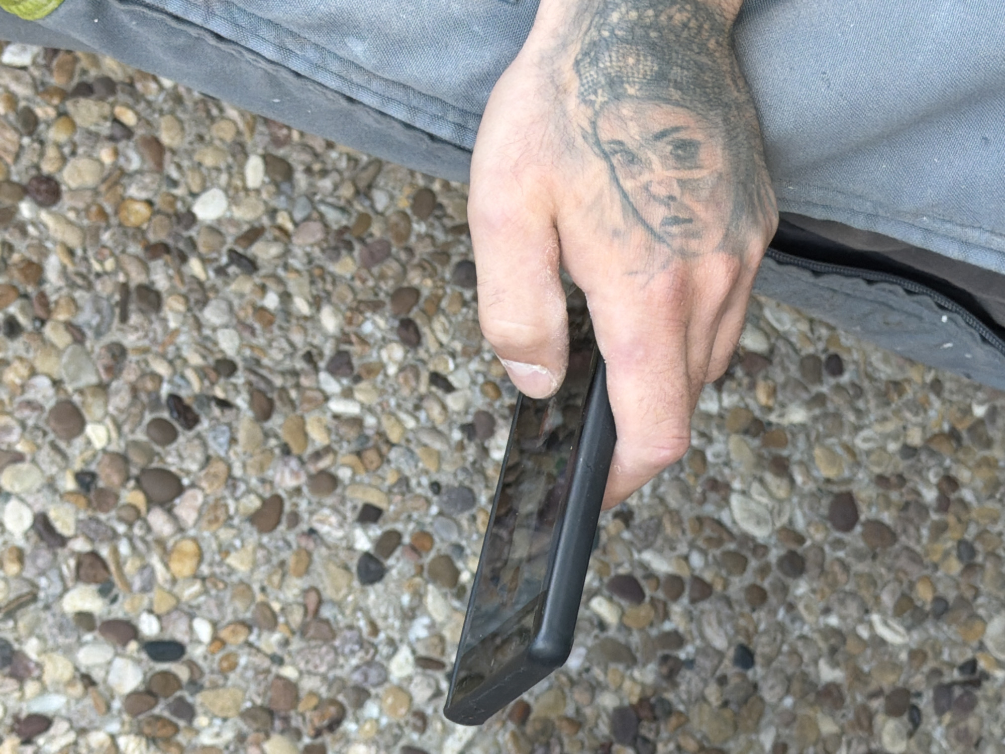 A close-up side view shows a person's tattooed hand holding a black smartphone. The person is wearing grey work overalls. The background is a textured wall made of small, multicolored pebbles. The scene appears to be outdoors, with the lighting suggesting daytime. The location is in Leudelange, Luxembourg.