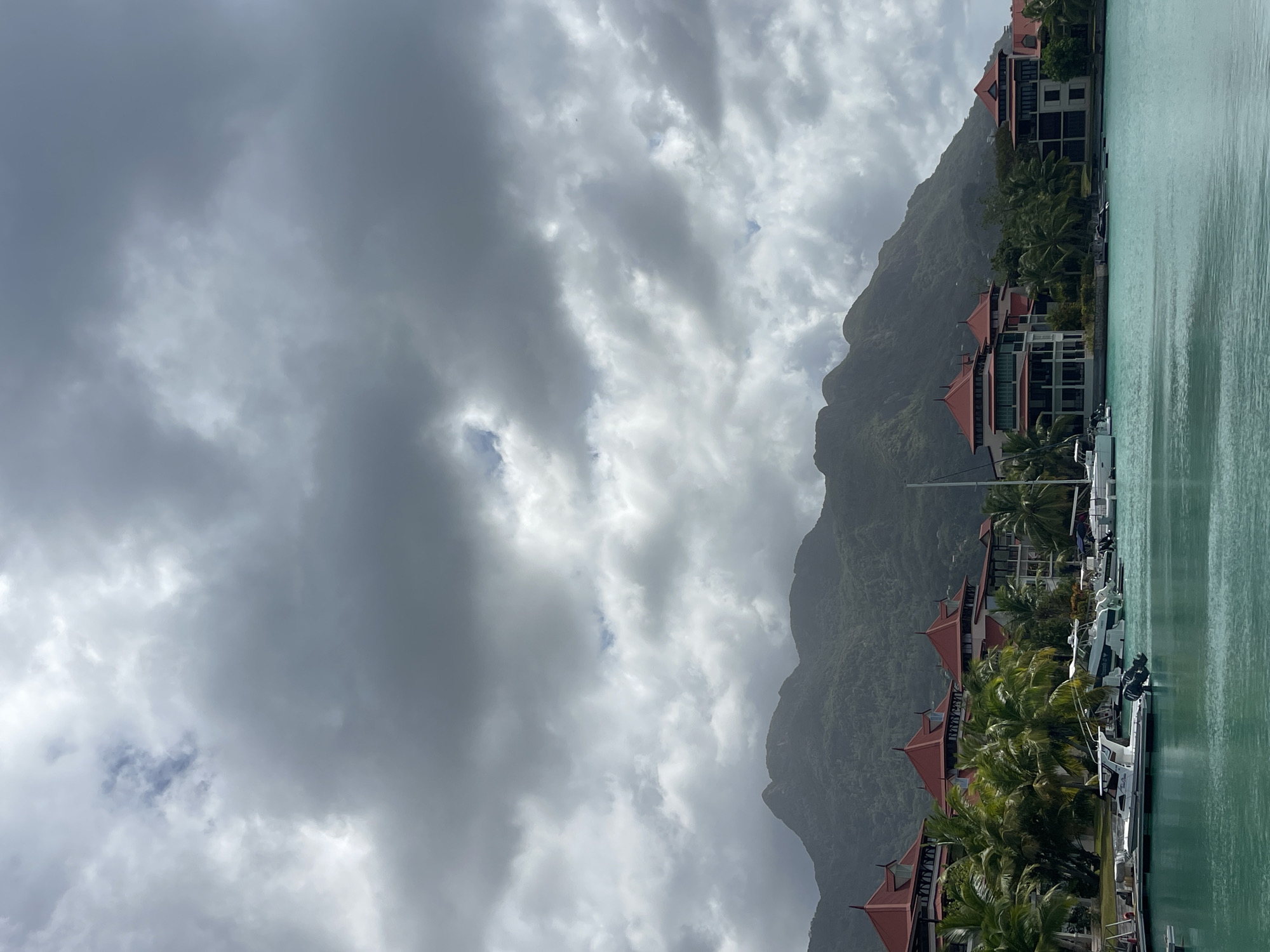 A wide-angle, vertical shot captures a coastal resort with a harbor in the foreground, mountains in the background, and a cloudy sky above. The water in the harbor is a vibrant turquoise color. Several white boats are docked, including speedboats and a sailboat.
The resort buildings are visible behind the harbor. They feature distinct red-tiled roofs and white or cream-colored facades with multiple stories. Palm trees are interspersed among the buildings and along the waterfront.
The mountains in the background are covered in lush green vegetation and appear mist-covered due to the cloudy sky. The sky itself is filled with heavy, dark gray clouds, with some lighter patches suggesting potential breaks in the overcast.
The scene depicts a tranquil, tropical resort environment, likely located in or near Providence, Seychelles, given the presence of tropical foliage and mountainous terrain. There are no people visible in the image.