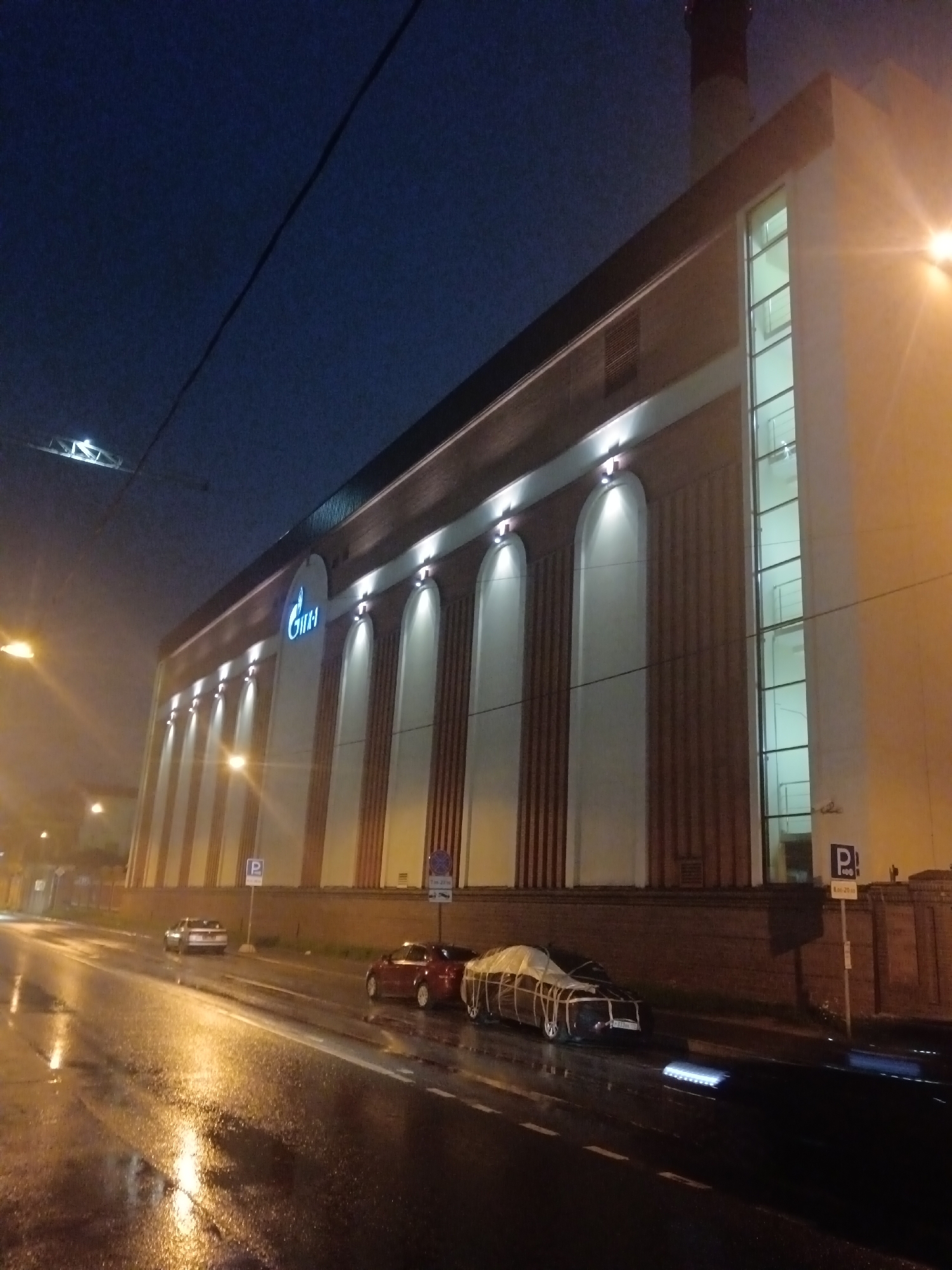 The image depicts a night scene in front of a modern building with a distinctive Gazprom logo illuminated on its facade. The building features a facade with vertical, arched indentations and a prominent glass section on the right side. Several streetlights cast a warm glow on the wet pavement, reflecting the light from the building and street lamps. Cars are parked along the street, and one car in the foreground is covered with a white tarp. The overall atmosphere is somewhat moody and urban, with the dark sky and wet road suggesting recent rain. The presence of the Gazprom logo indicates the location is likely a corporate building or office in Saint Petersburg, Russia.